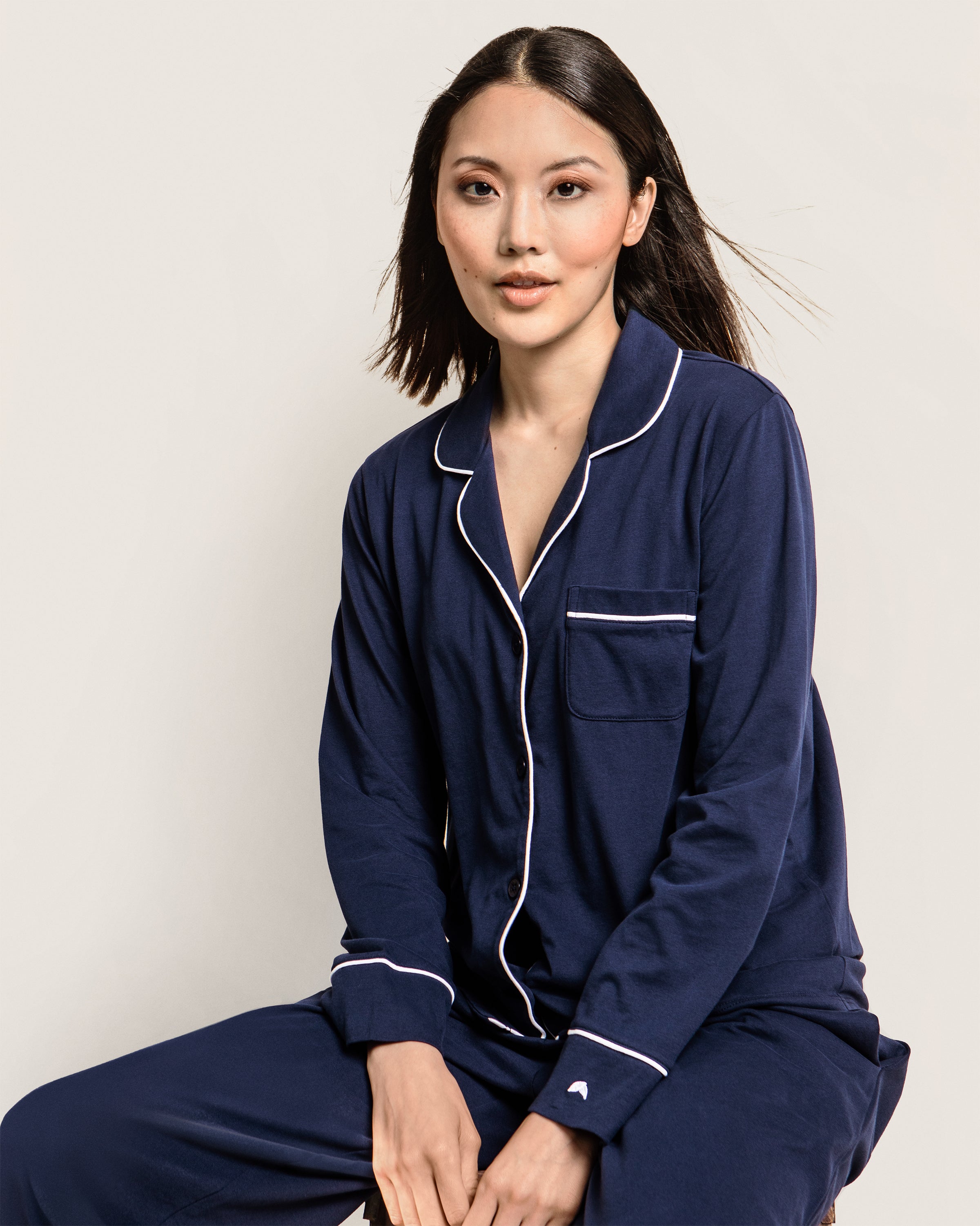 A person in Petite Plumes Women’s Pima Pajama Set in Navy sits against a light backdrop. The luxurious sleepwear, made of soft Pima cotton, showcases white piping, a front pocket, and button-down details. Their long hair blows gently to the side for an ethereal effect.