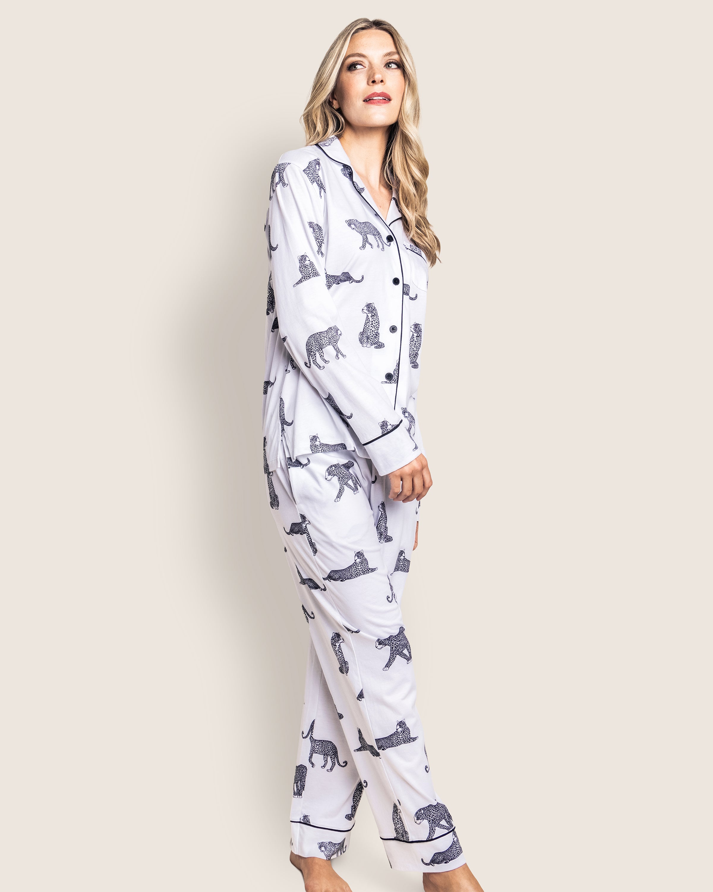 A woman stands wearing the Womens Pima Pajama Set in Panthère de Paris by Petite Plume, crafted from luxe Peruvian Pima cotton with black animal prints. She gazes upward with a neutral expression against a plain backdrop.