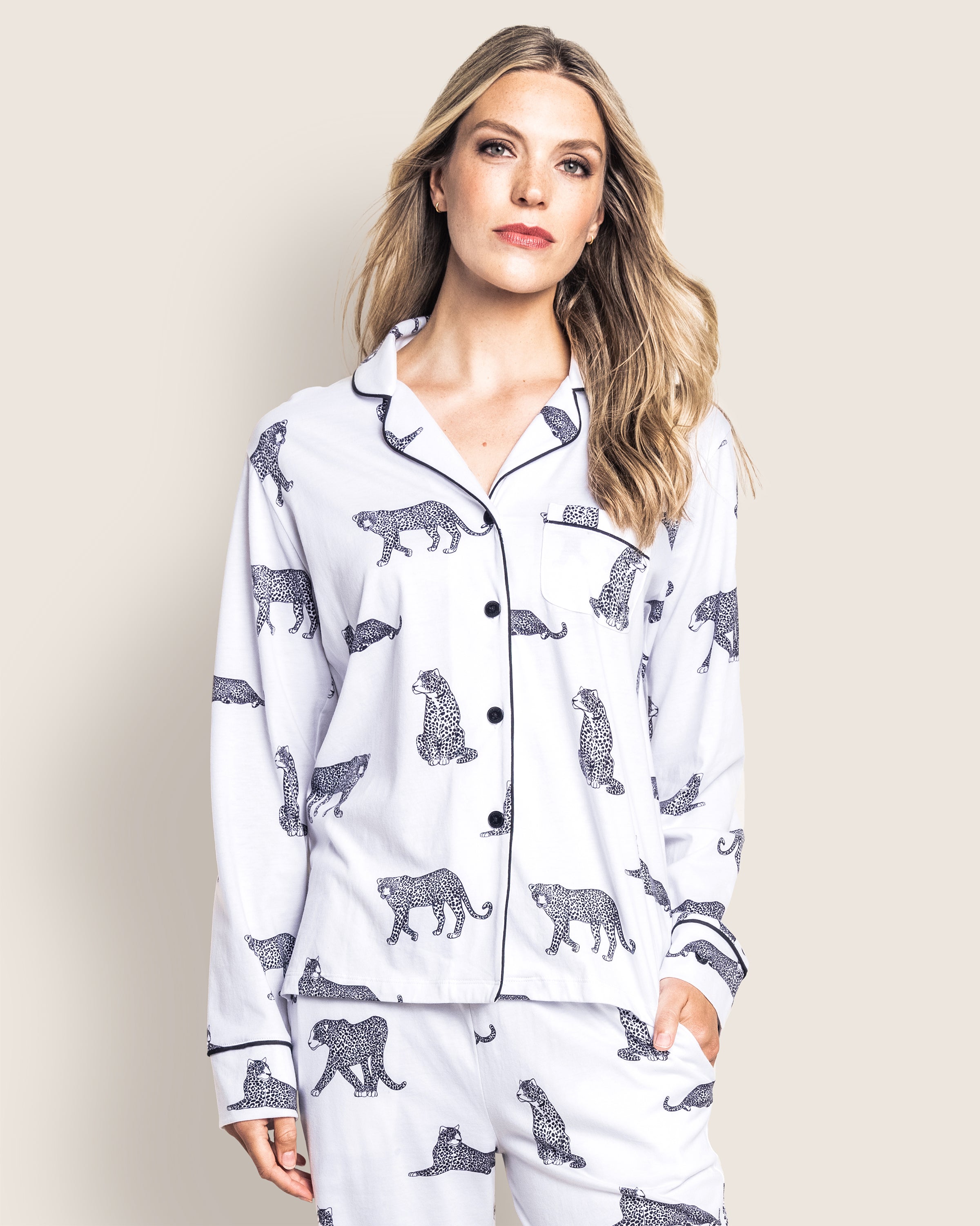 A woman with long blonde hair wears the elegant Womens Pima Pajama Set in Panthère de Paris by Petite Plume, featuring a striking black leopard print on white Luxe Pima cotton. She stands with one hand in her pocket against a plain, light background.
