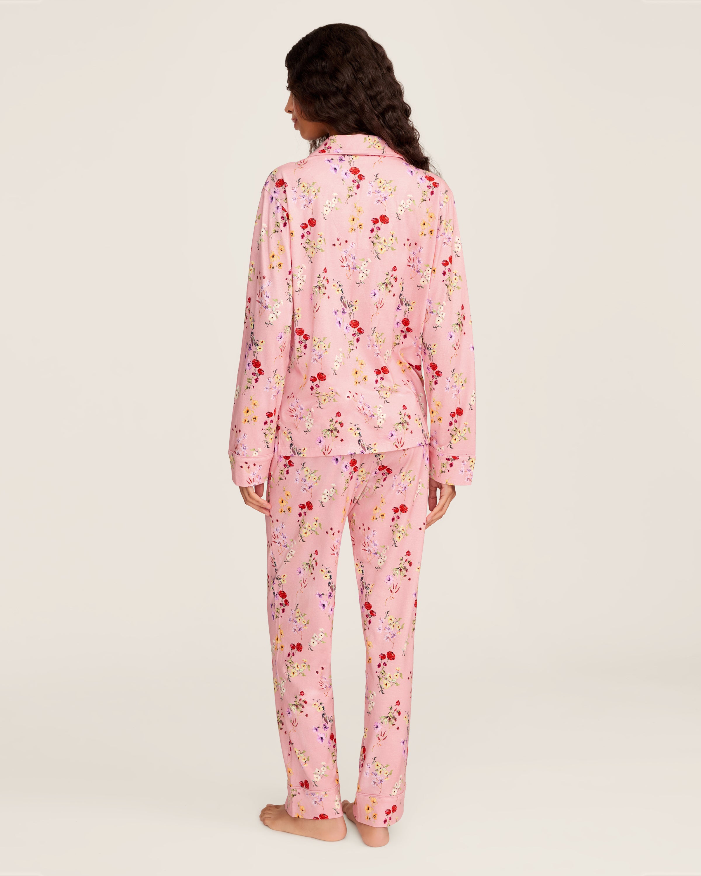The Women's Pima Pajama Set in Brilliant Botanical Blush features a pink button-up long-sleeve top and matching pants, both adorned with a vibrant floral print in red, white, and purple.