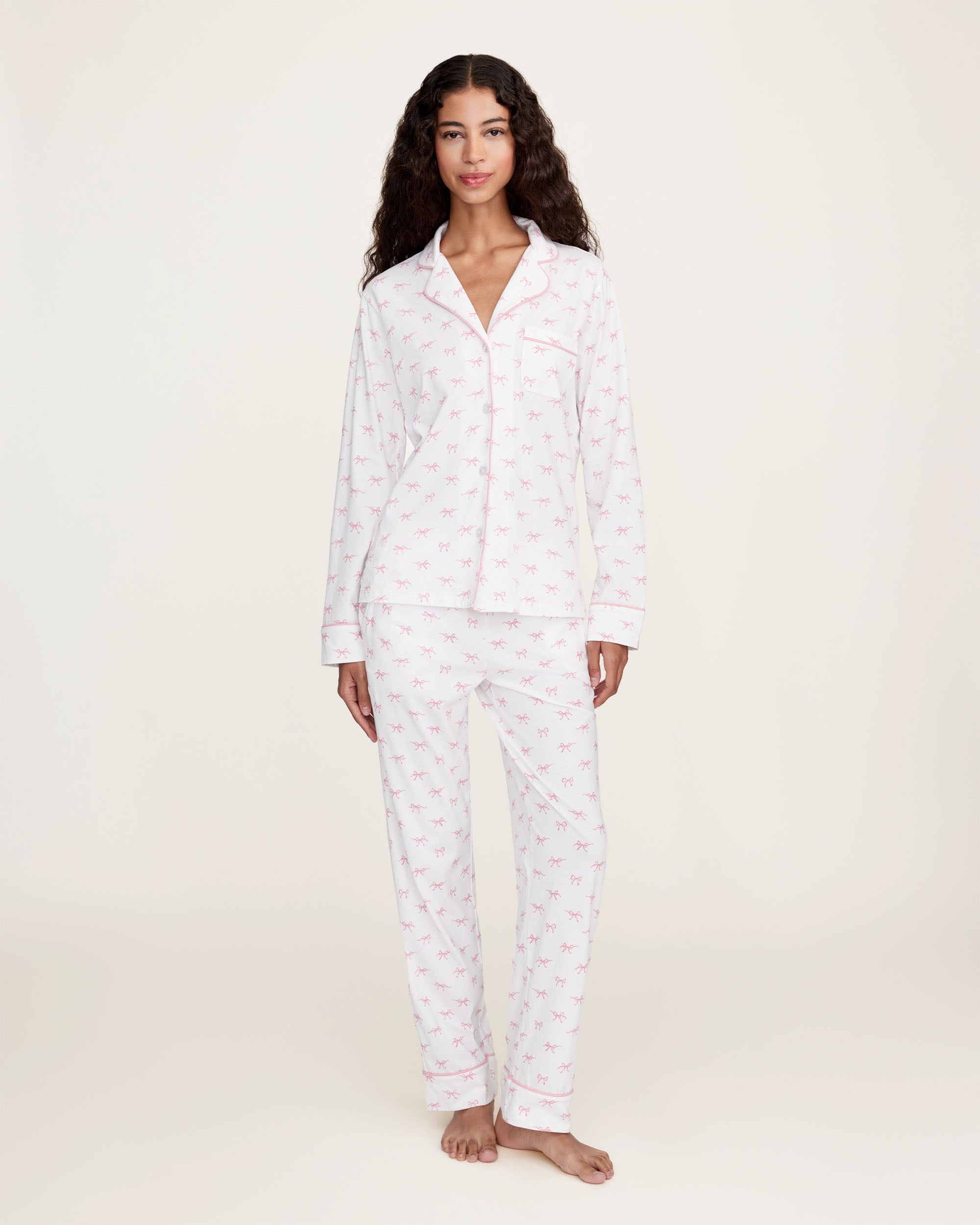 The Women's Pima Pajama Set in Blushing Bows features a long-sleeve button-up top and pants in white fabric with a small pink bow pattern and pink piping accents.