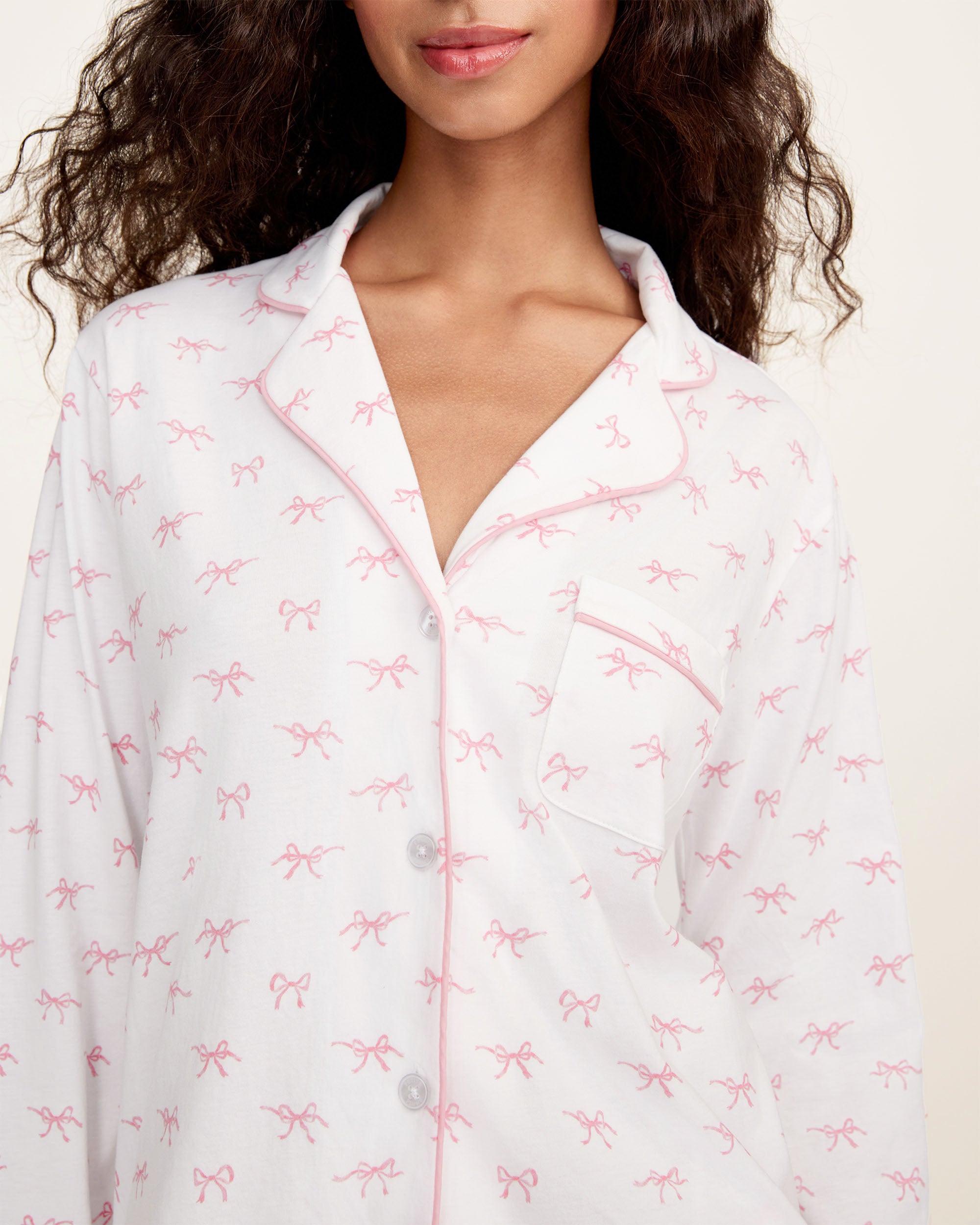 The Women's Pima Pajama Set in Blushing Bows features a long-sleeve button-up top and pants in white fabric with a small pink bow pattern and pink piping accents.
