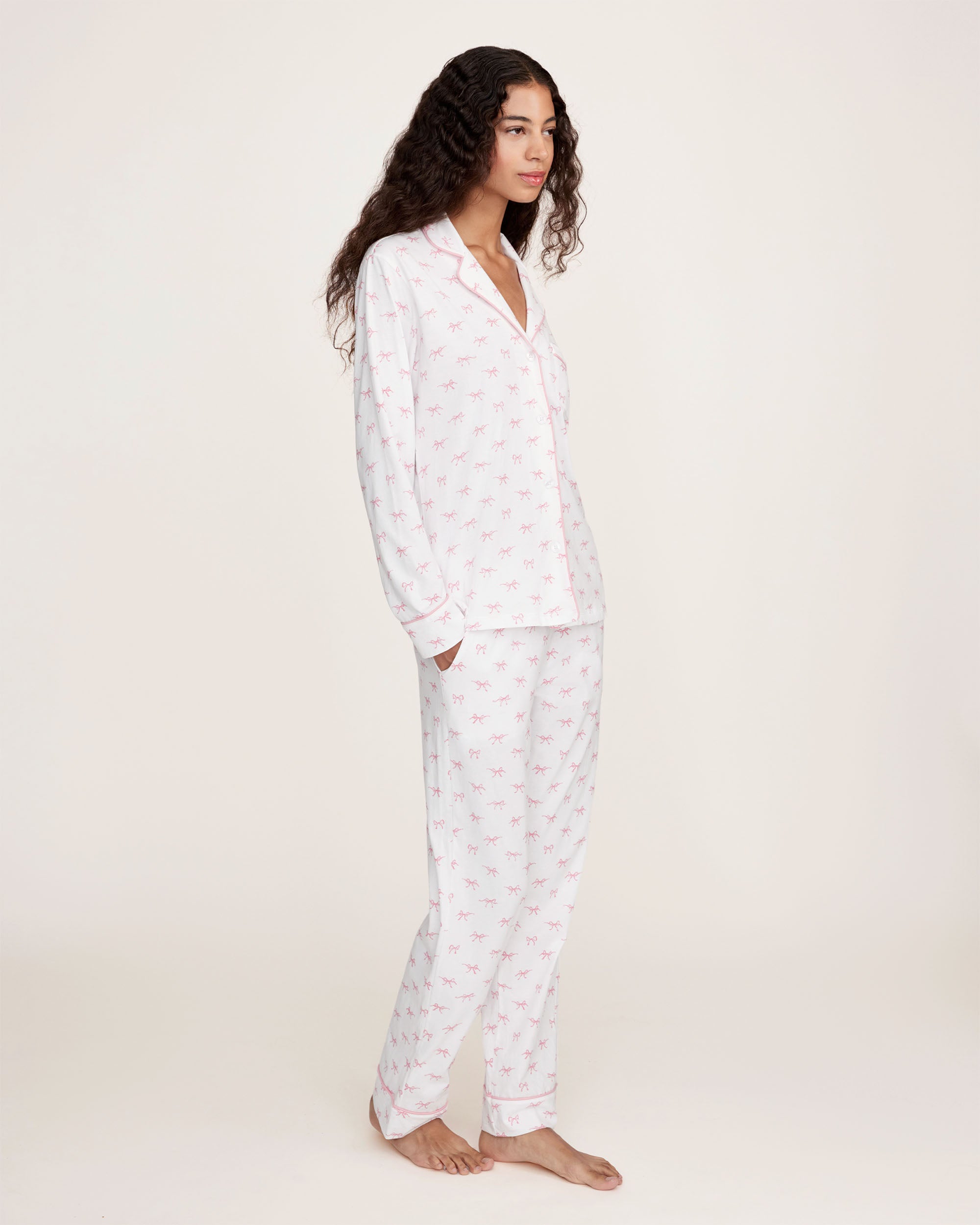 The Women's Pima Pajama Set in Blushing Bows features a long-sleeve button-up top and pants in white fabric with a small pink bow pattern and pink piping accents.
