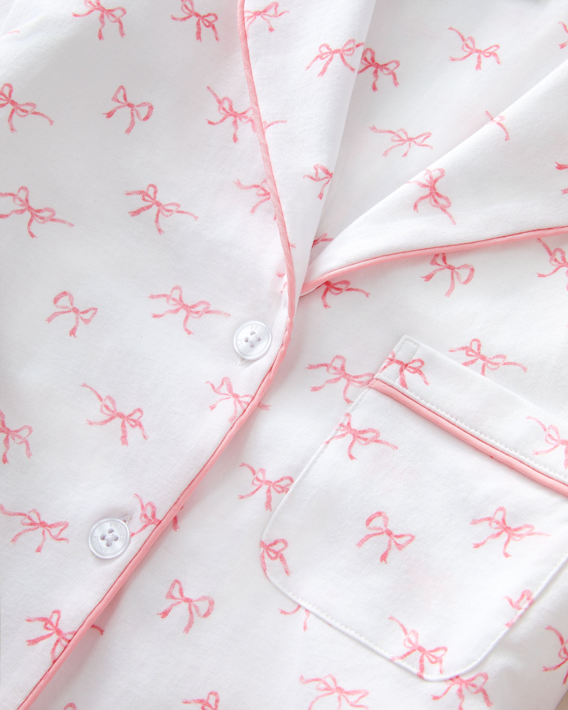 The Women's Pima Pajama Set in Blushing Bows features a long-sleeve button-up top and pants in white fabric with a small pink bow pattern and pink piping accents.