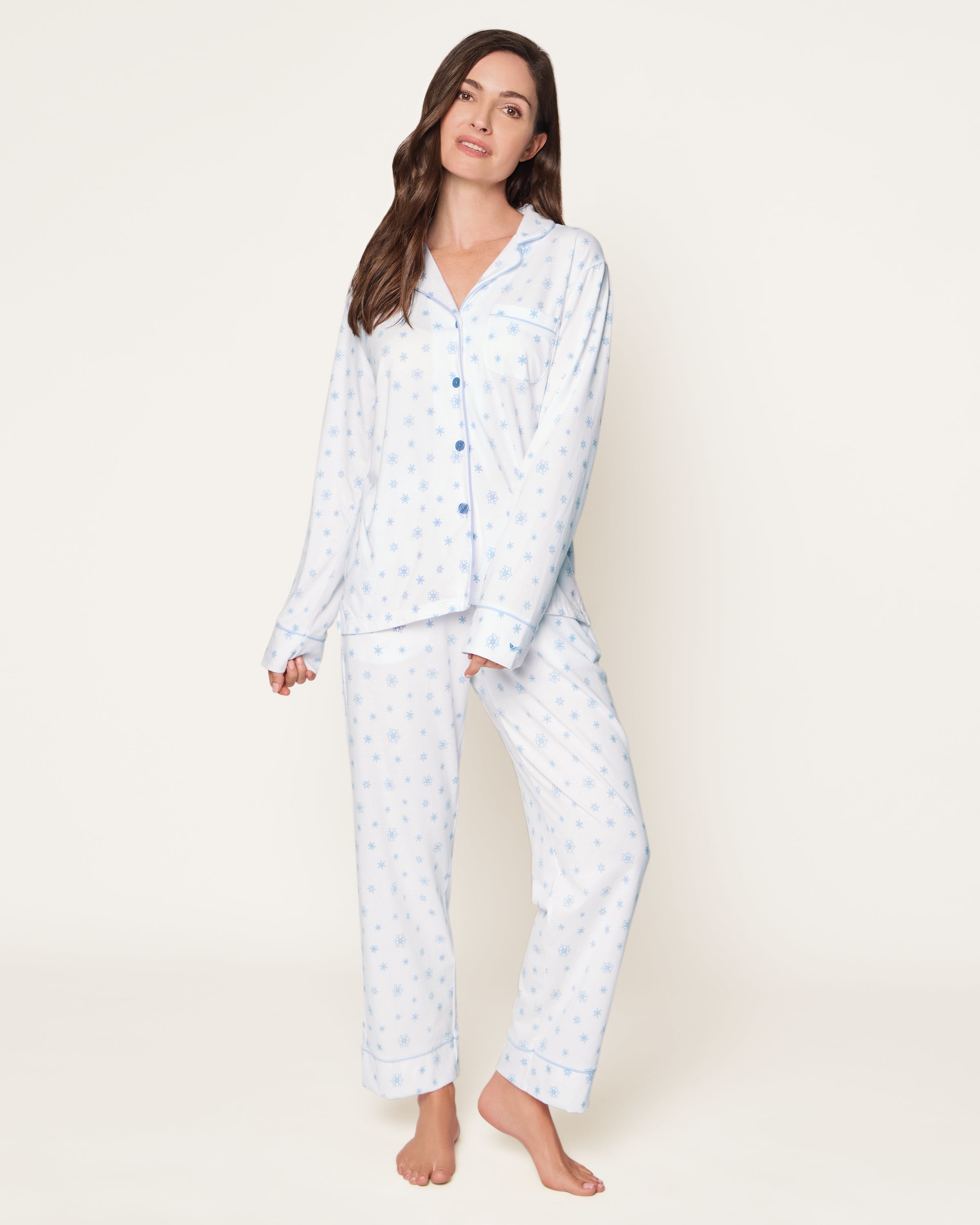 The Women's Pima Pajama Set in Periwinkle Wonderland features white pajamas with light blue snowflake patterns, a collared long-sleeve button-up top with pocket, and matching elastic waist pants, both accented with blue piping.