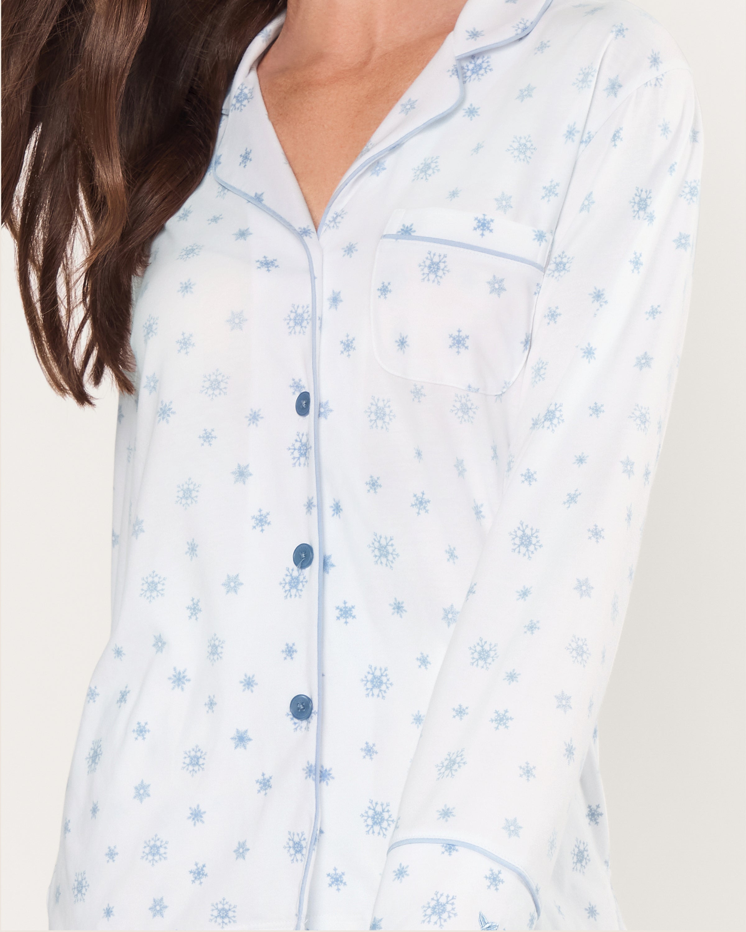 The Women's Pima Pajama Set in Periwinkle Wonderland features white pajamas with light blue snowflake patterns, a collared long-sleeve button-up top with pocket, and matching elastic waist pants, both accented with blue piping.