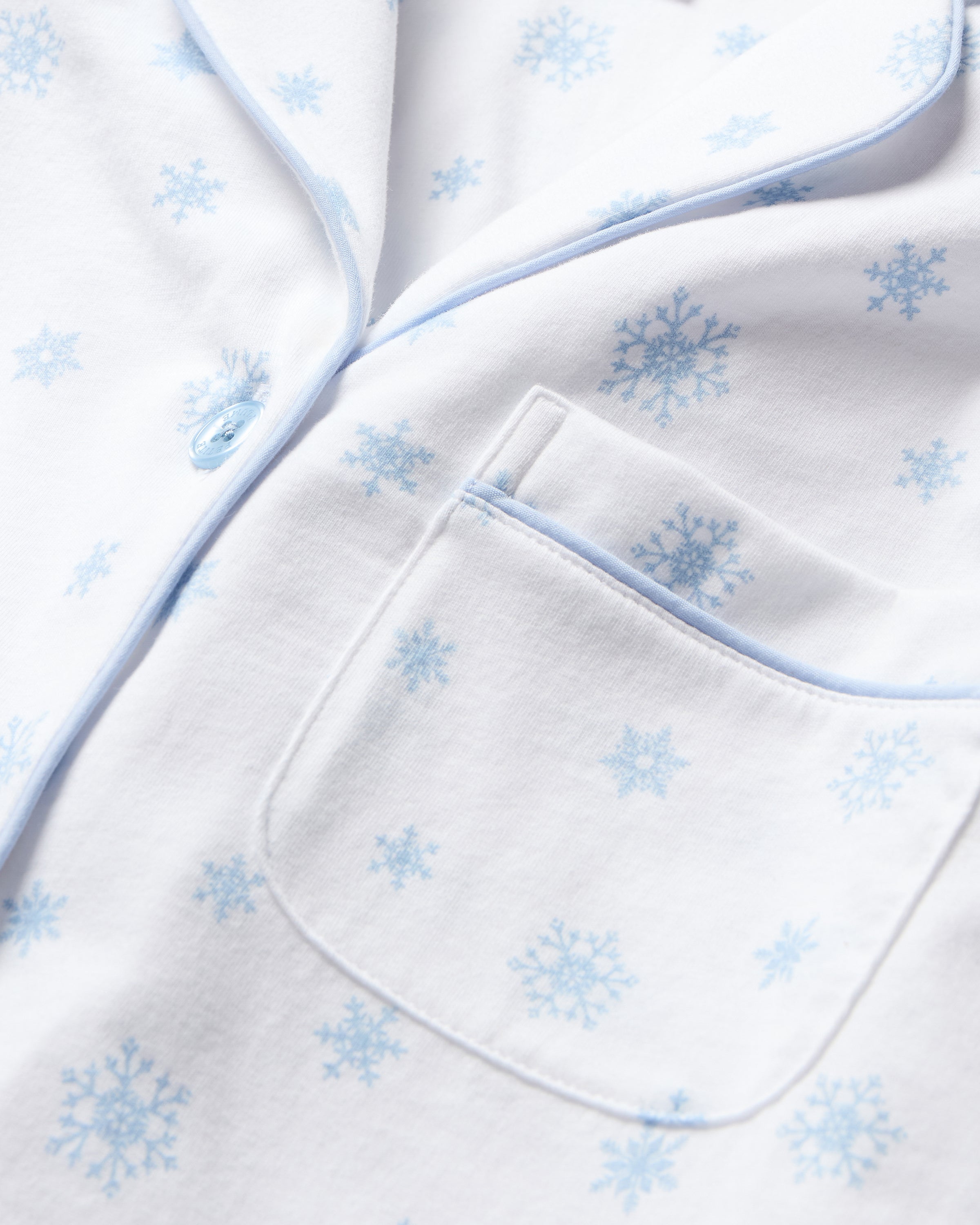 The Women's Pima Pajama Set in Periwinkle Wonderland features white pajamas with light blue snowflake patterns, a collared long-sleeve button-up top with pocket, and matching elastic waist pants, both accented with blue piping.