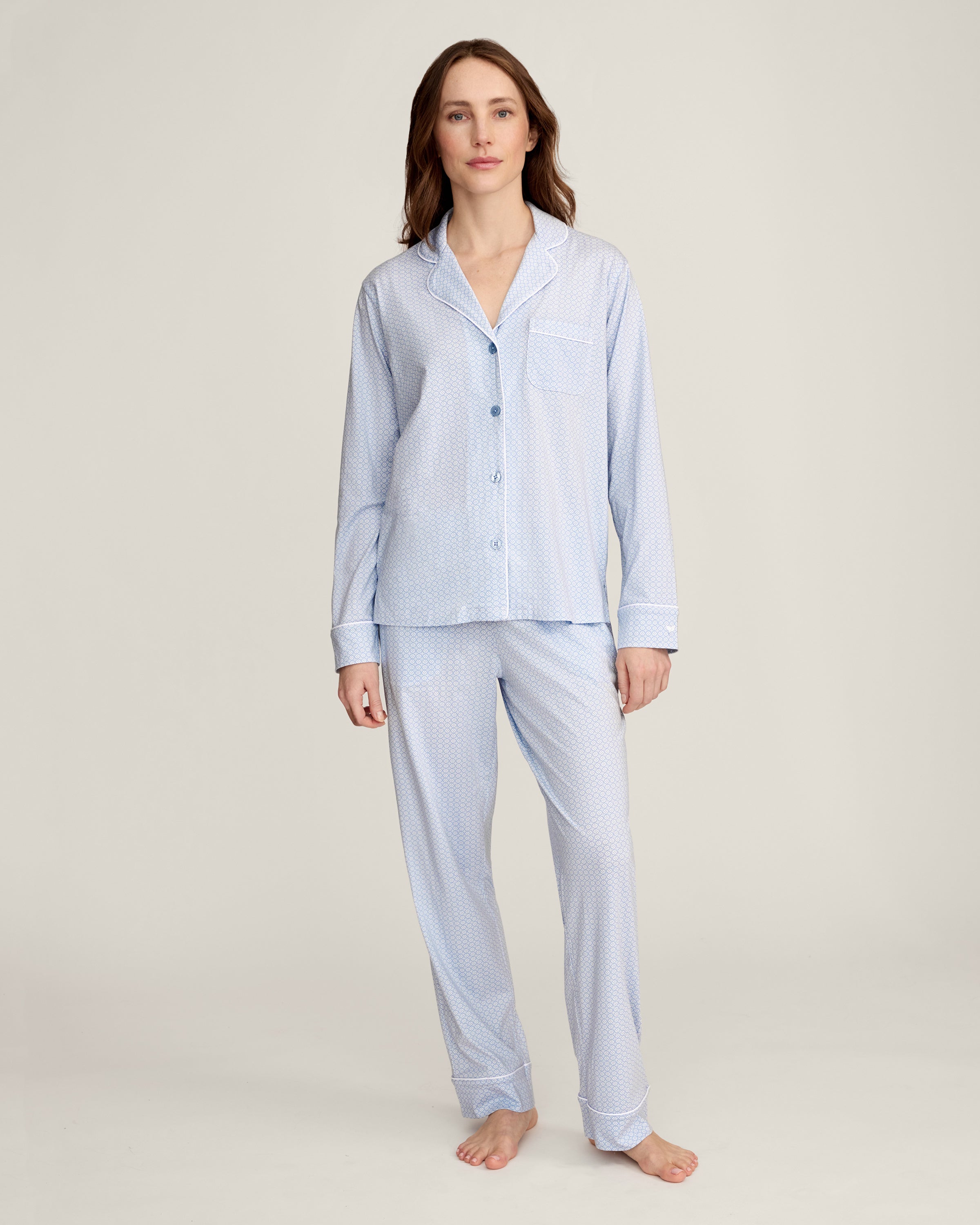 A woman with light brown hair and a neutral expression stands barefoot against a plain background, modeling the Women's Lightweight Pima Pajama Set in Periwinkle Geo—featuring long sleeves, button-up top with collar, and matching pants.