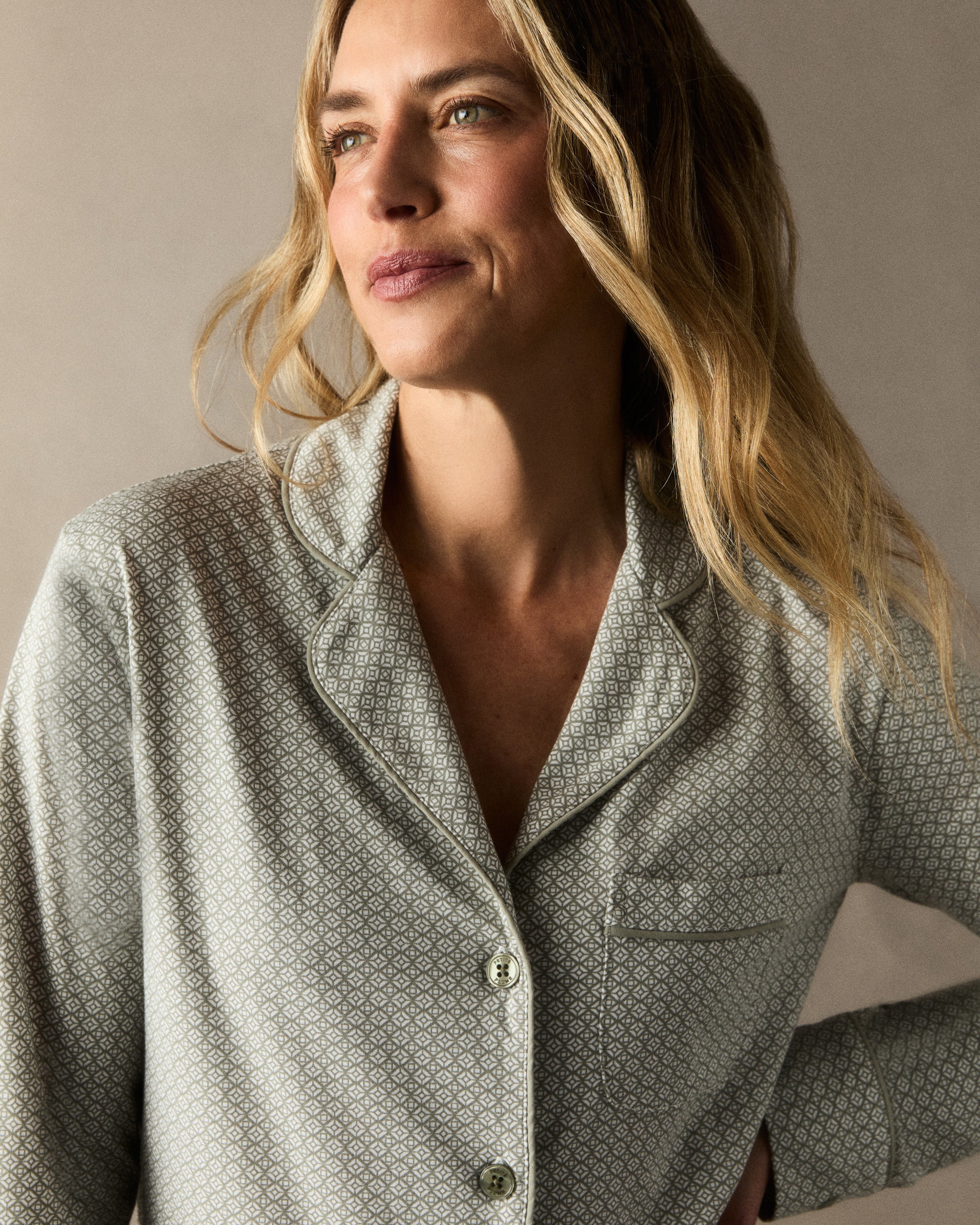 A woman with long curly hair stands barefoot against a white background, wearing the Women's Lightweight Pima Pajama Set in Sage Geo—a light gray button-up long-sleeve top and matching pants with a small geometric pattern.