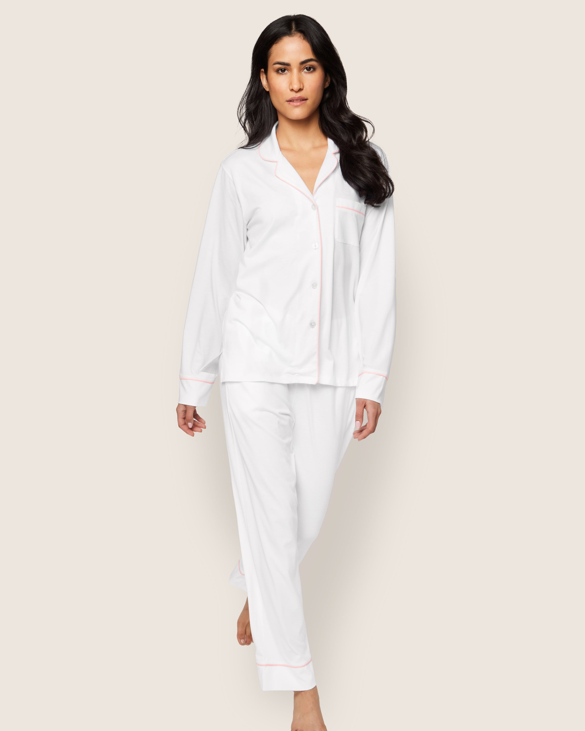 A person with long dark hair walks forward wearing the luxurious Womens Pima Pajama Set in White with Pink Piping by Petite Plume, set against an off-white background.