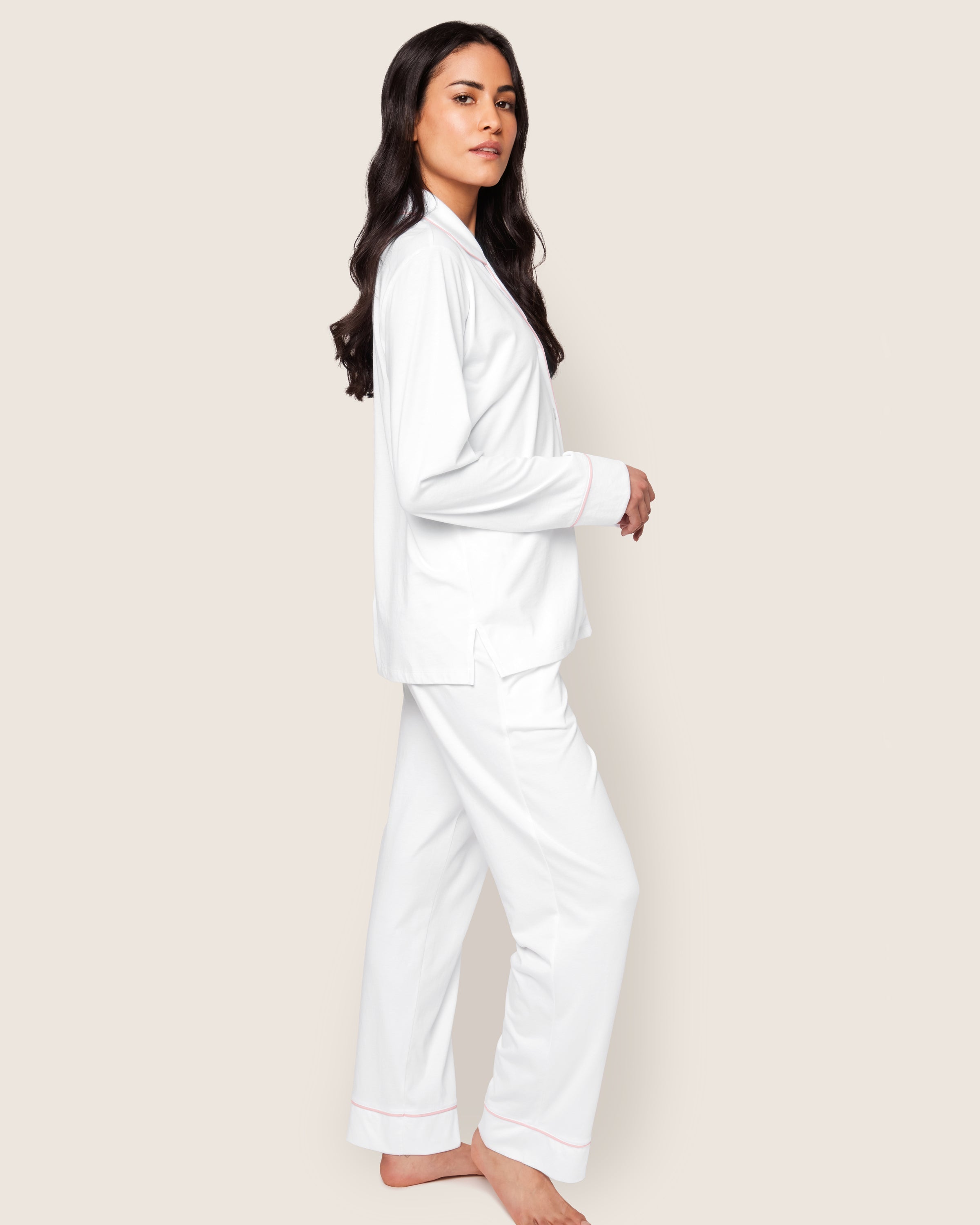 A person with long dark hair is standing sideways, barefoot against a light background, wearing Petite Plume’s luxury Womens Pima Pajama Set in White with Pink Piping.