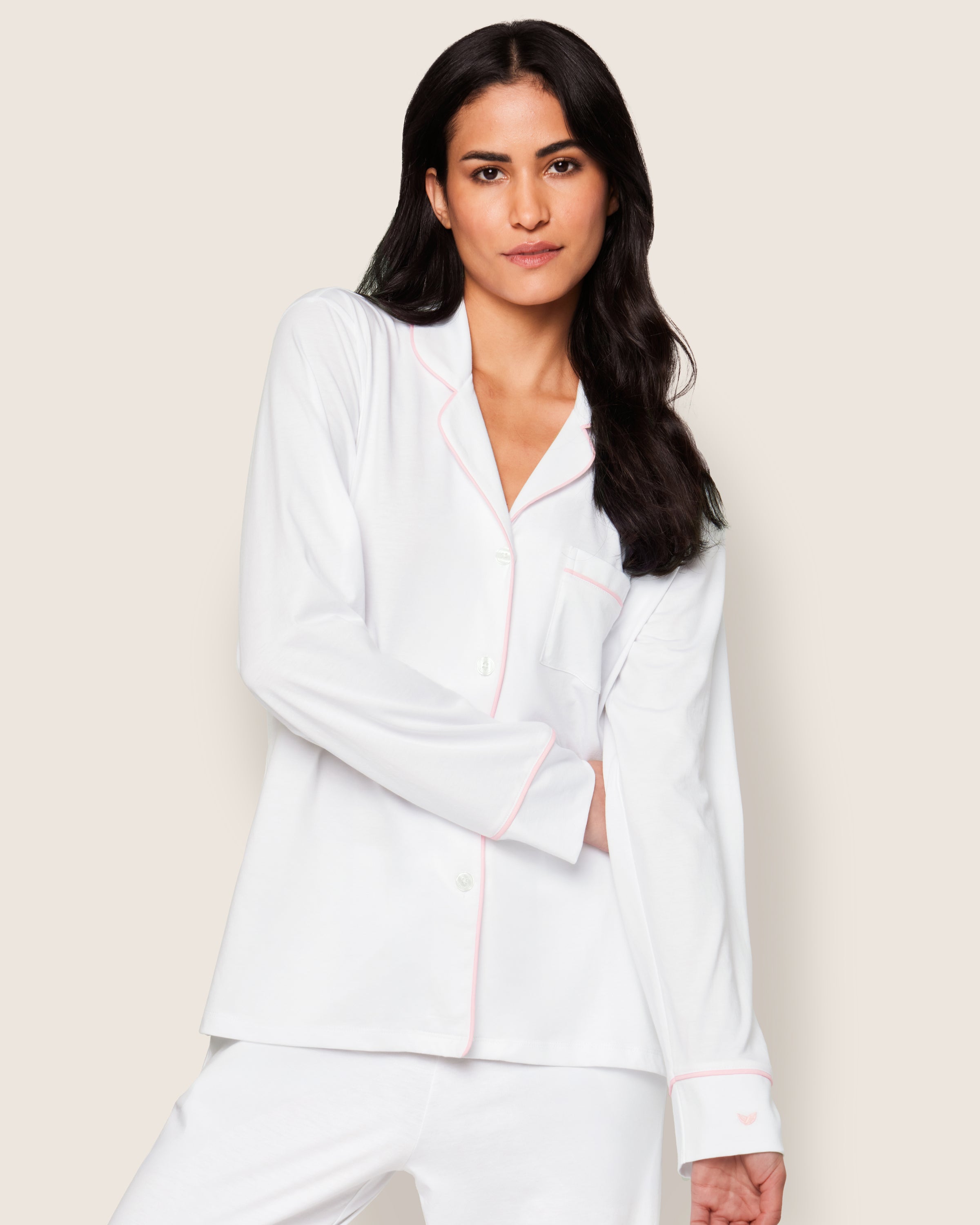 A person with long dark hair is wearing the luxurious Womens Pima Pajama Set in White with Pink Piping by Petite Plume. They stand with arms crossed and a neutral expression against an off-white background.