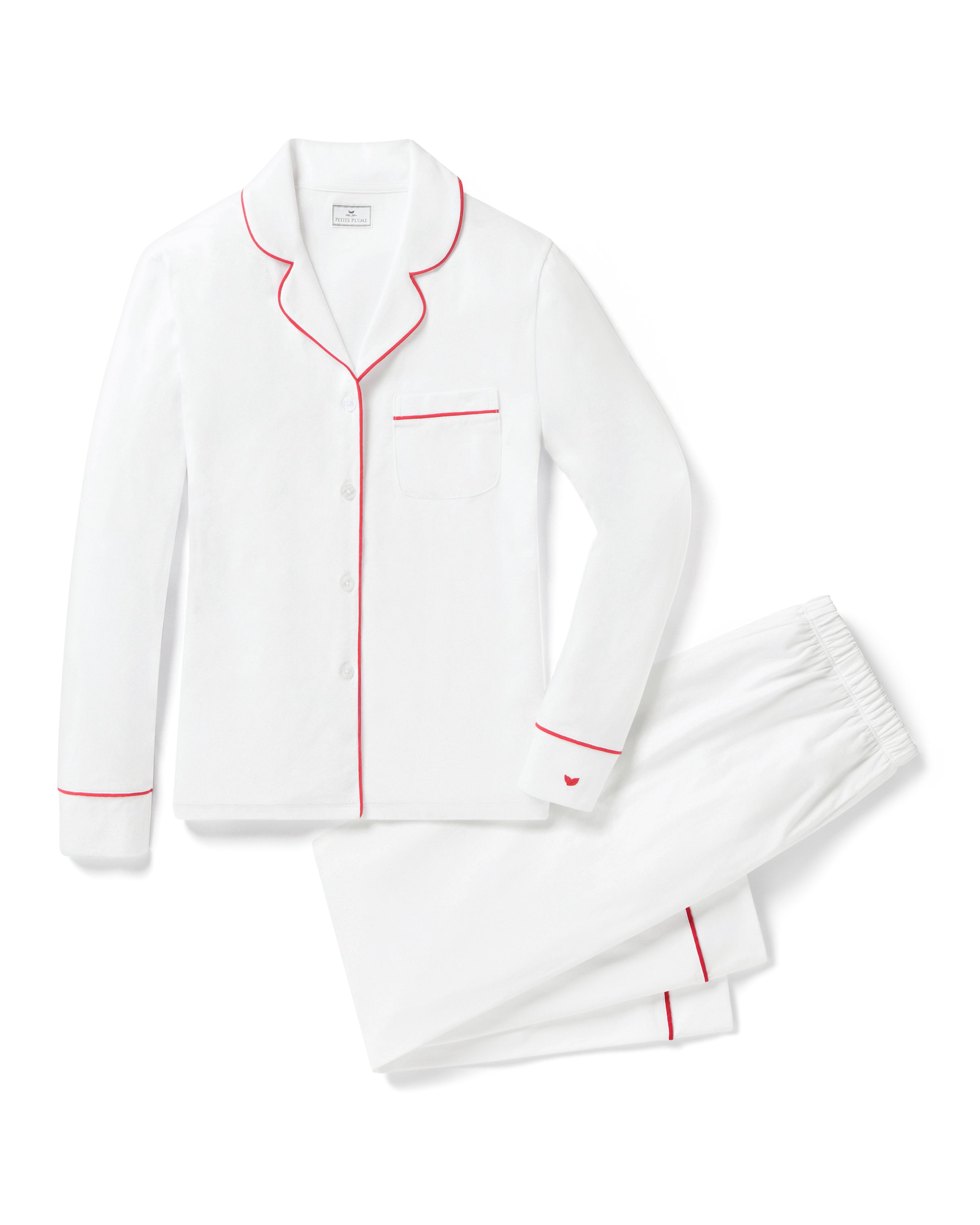 A person stands barefoot against a plain background in a Womens White Pima Pajama Set with Red Piping by Petite Plume, crafted from Peruvian Pima cotton. Their curly hair and classic pajama silhouette exude elegance as they smile with hands in pockets.