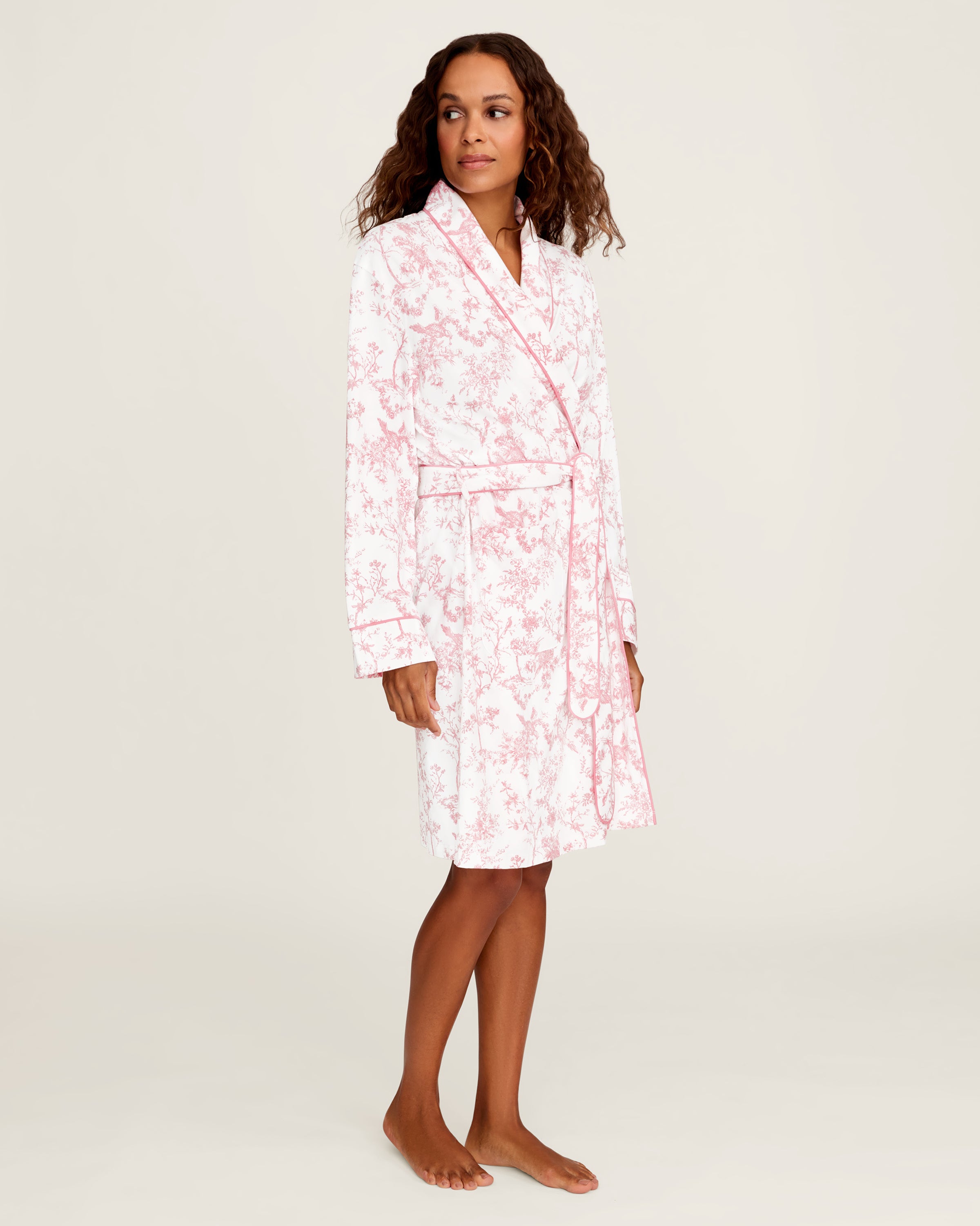 The Women's Pima Robe in Pink Timeless Toile features a white knee-length design with a pink floral toile pattern, long sleeves, a shawl collar, and a matching waist belt.