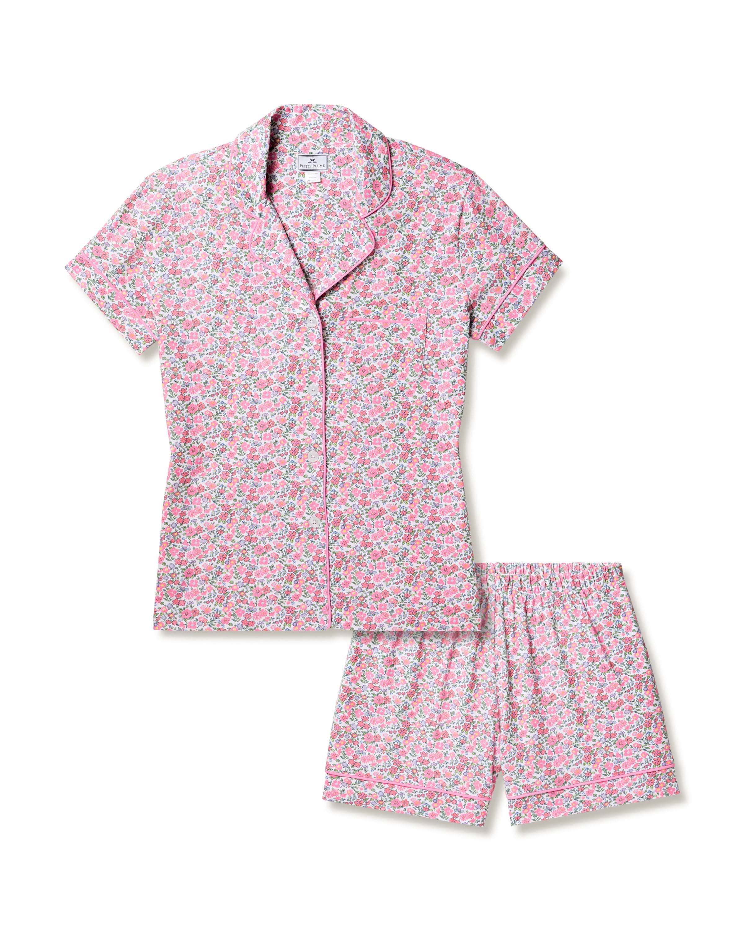 The Womens Pima Short Sleeve Short Set in Fleurs de Rose by Petite Plume offers a button-up top and shorts. Its fabric evokes a summer garden vibe with a light pink background and colorful floral patterns in pink, green, and white.