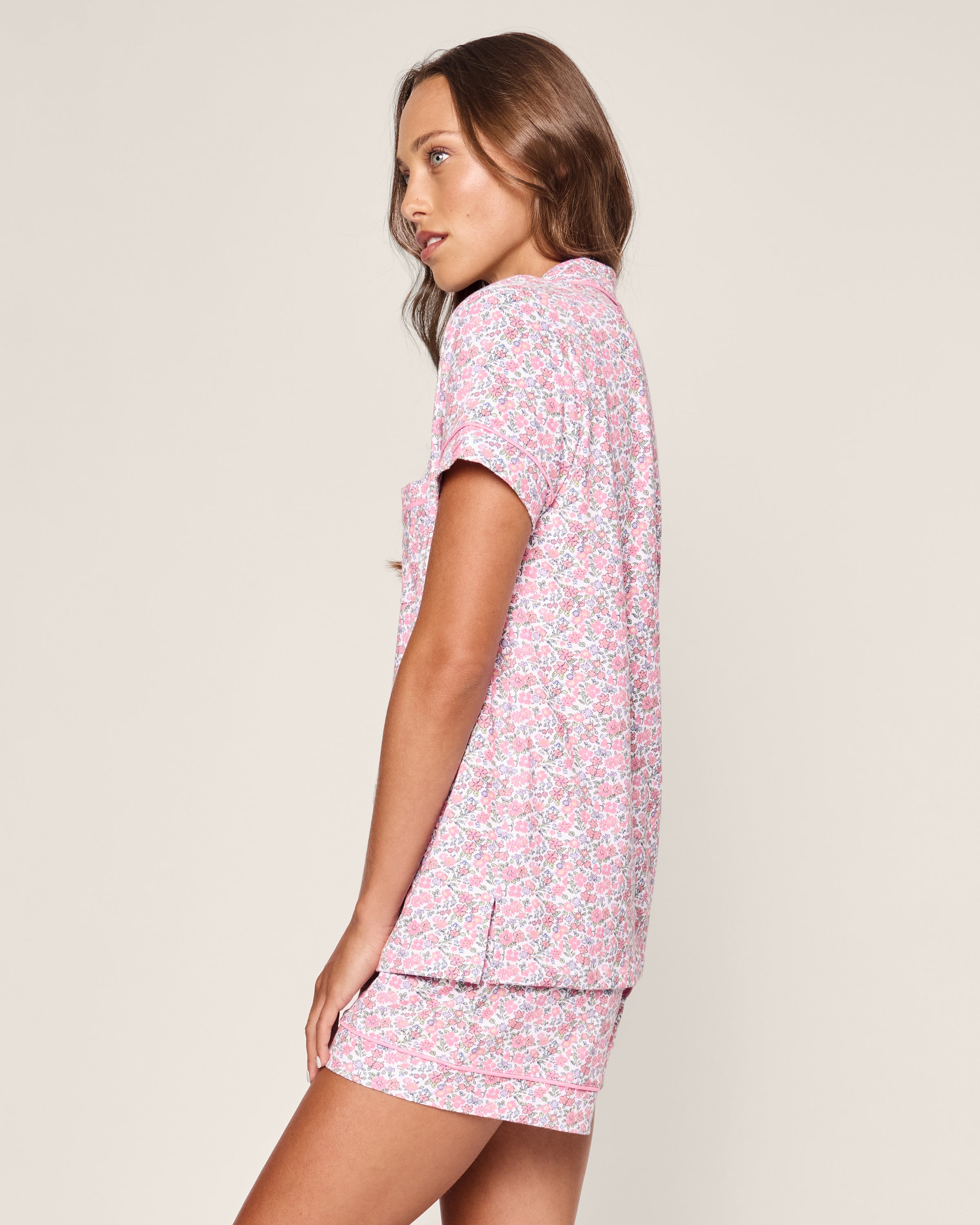 A woman stands sideways wearing the Womens Pima Short Sleeve Short Set in Fleurs de Rose by Petite Plume. The pink floral print exudes a breezy summer garden vibe with its collared top and relaxed-fit shorts, set against a plain backdrop highlighting the casual, summery look.