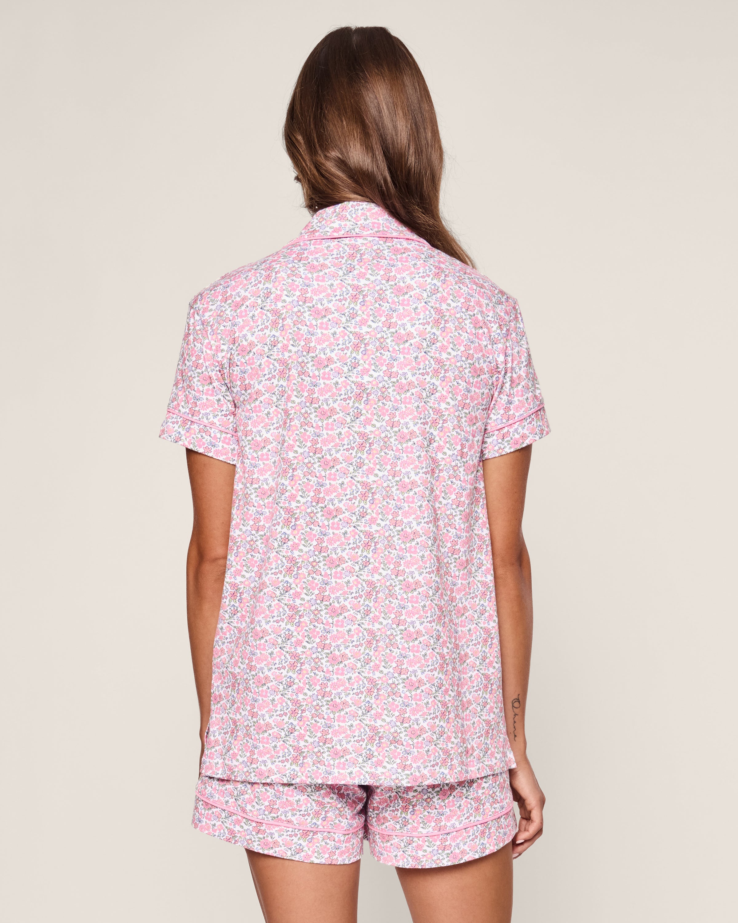 A person with long hair stands with their back to the camera, wearing Petite Plumes Womens Pima Short Sleeve Short Set in Fleurs de Rose, evoking a soothing garden charm against a plain background.