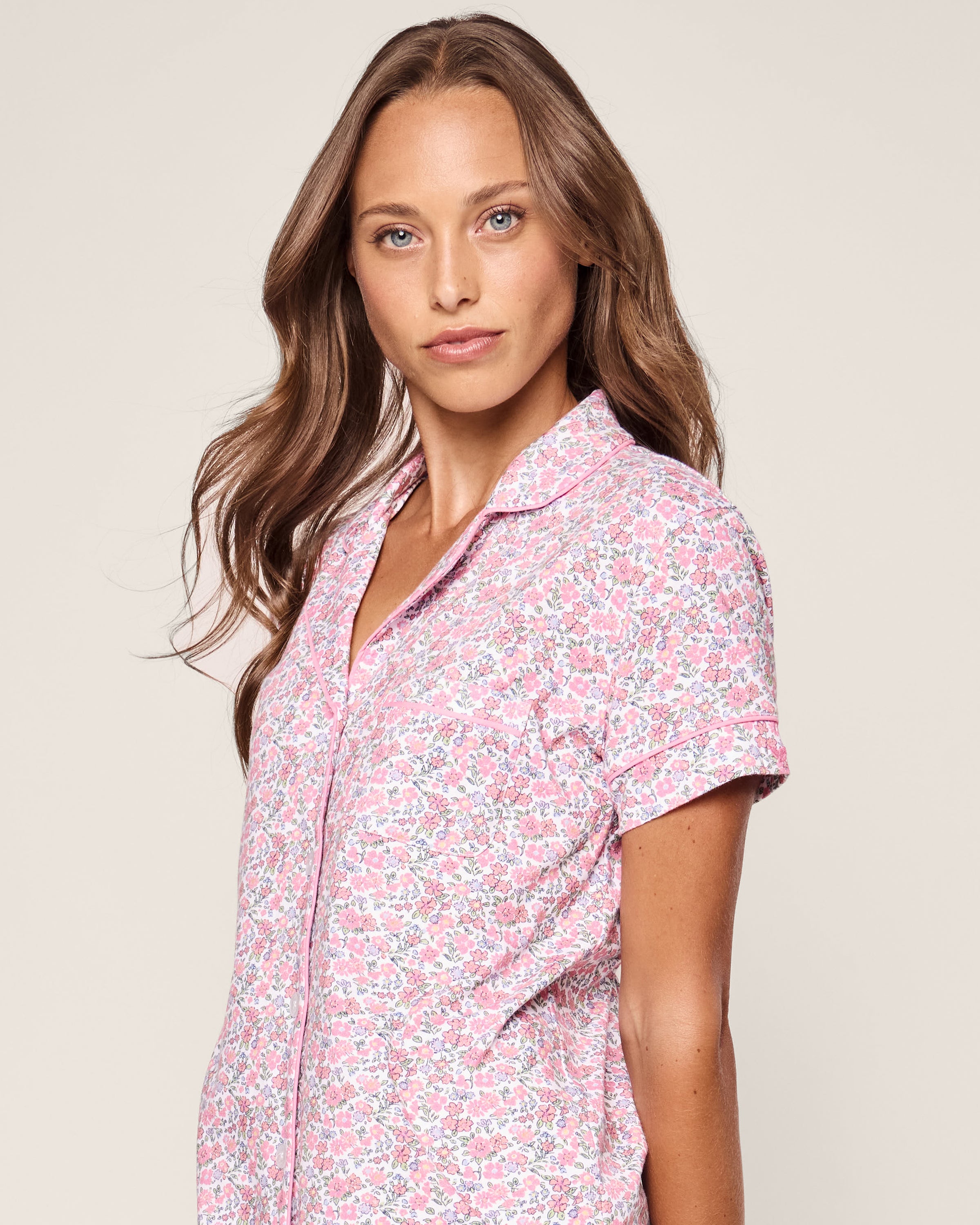 A woman with long brown hair and blue eyes wears the Womens Pima Short Sleeve Short Set in Fleurs de Rose from Petite Plume, gazing at the camera against a plain, light background that suggests a serene summer garden.