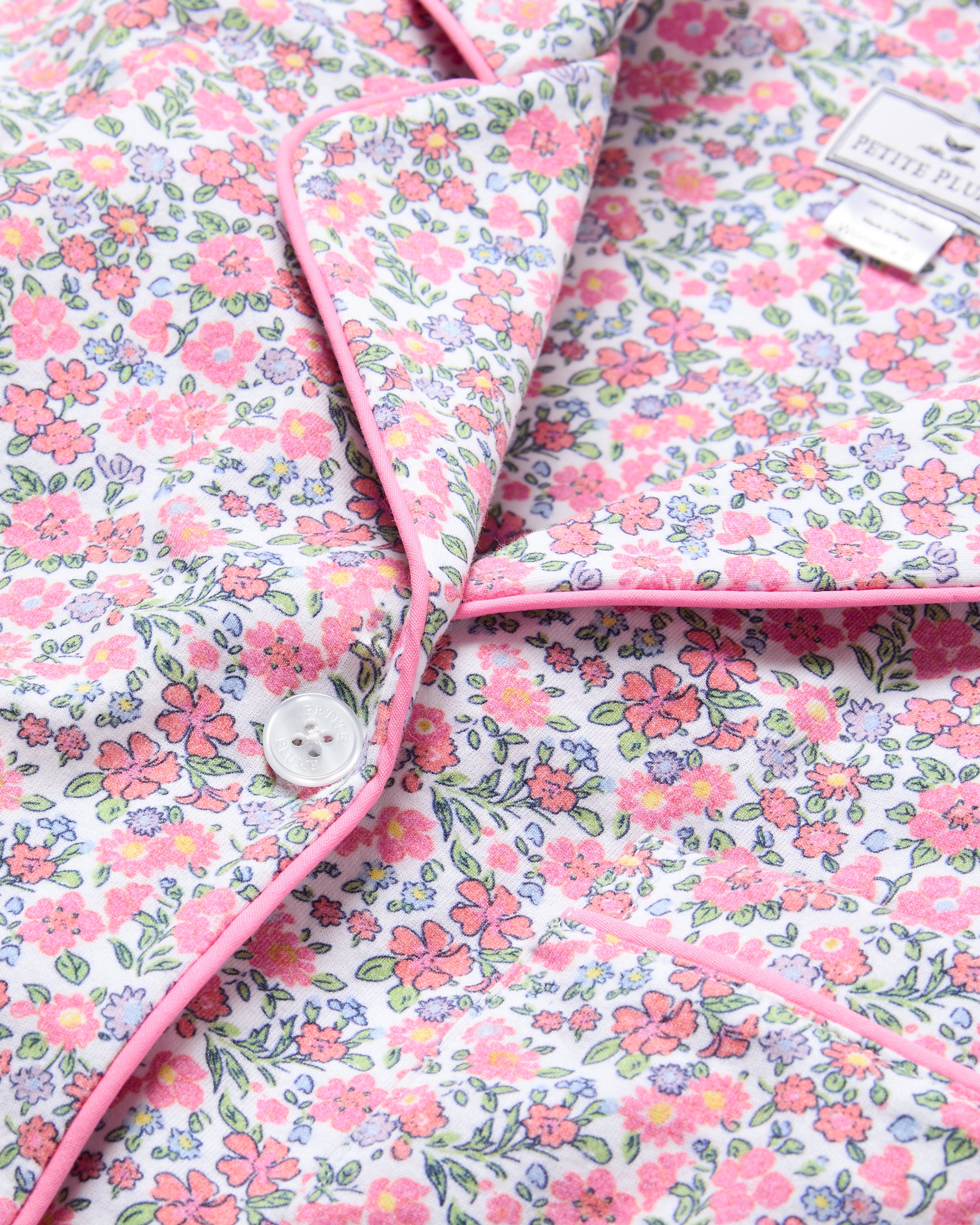 Close-up of a Womens Pima Short Sleeve Short Set in Fleurs de Rose by Petite Plume. This floral-patterned garment features pink and white flowers on a light background, pink piping, a white button, and a partially visible label at the upper right corner.