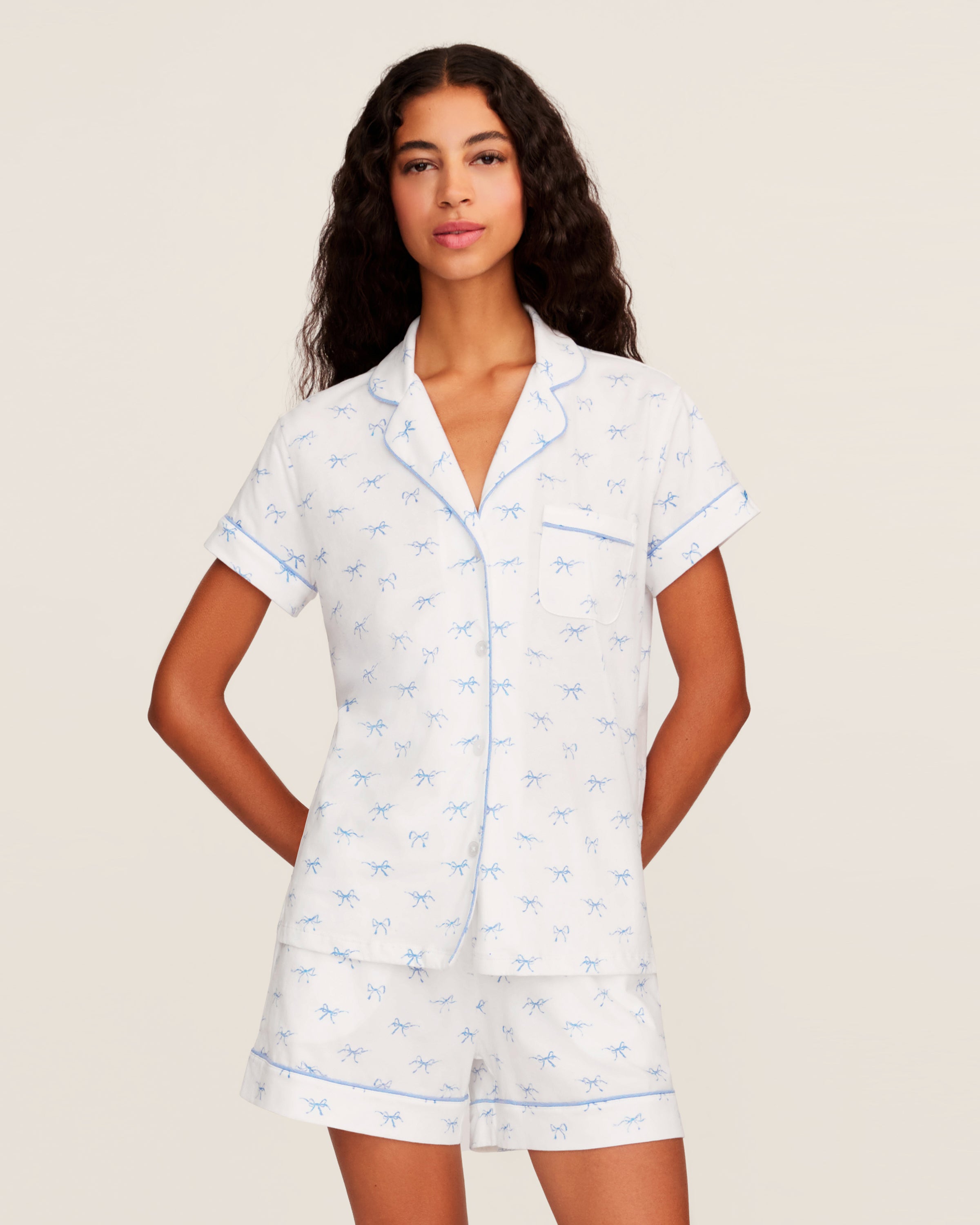 Women's Pima Short Sleeve Short Set in Something Blue Bows features a collared button-up top with chest pocket and matching elastic-waist shorts, both adorned with all-over blue bow print and contrast piping.