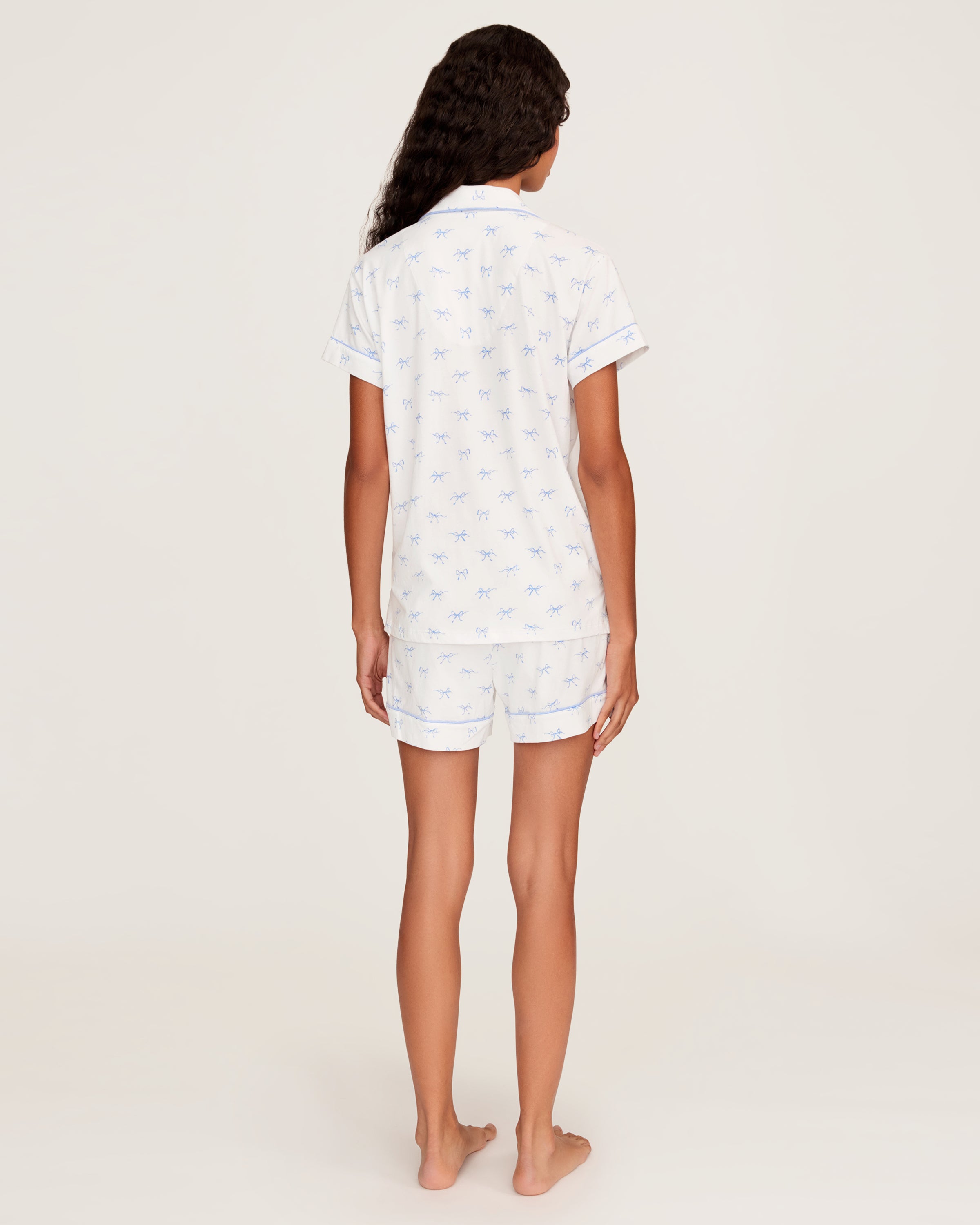Women's Pima Short Sleeve Short Set in Something Blue Bows features a collared button-up top with chest pocket and matching elastic-waist shorts, both adorned with all-over blue bow print and contrast piping.