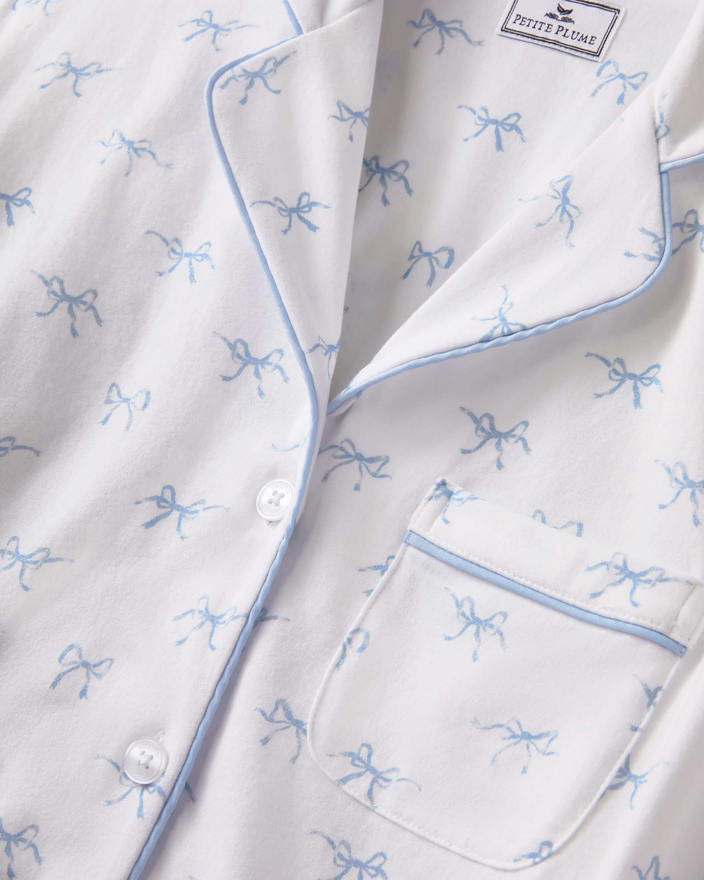 Women's Pima Short Sleeve Short Set in Something Blue Bows features a collared button-up top with chest pocket and matching elastic-waist shorts, both adorned with all-over blue bow print and contrast piping.