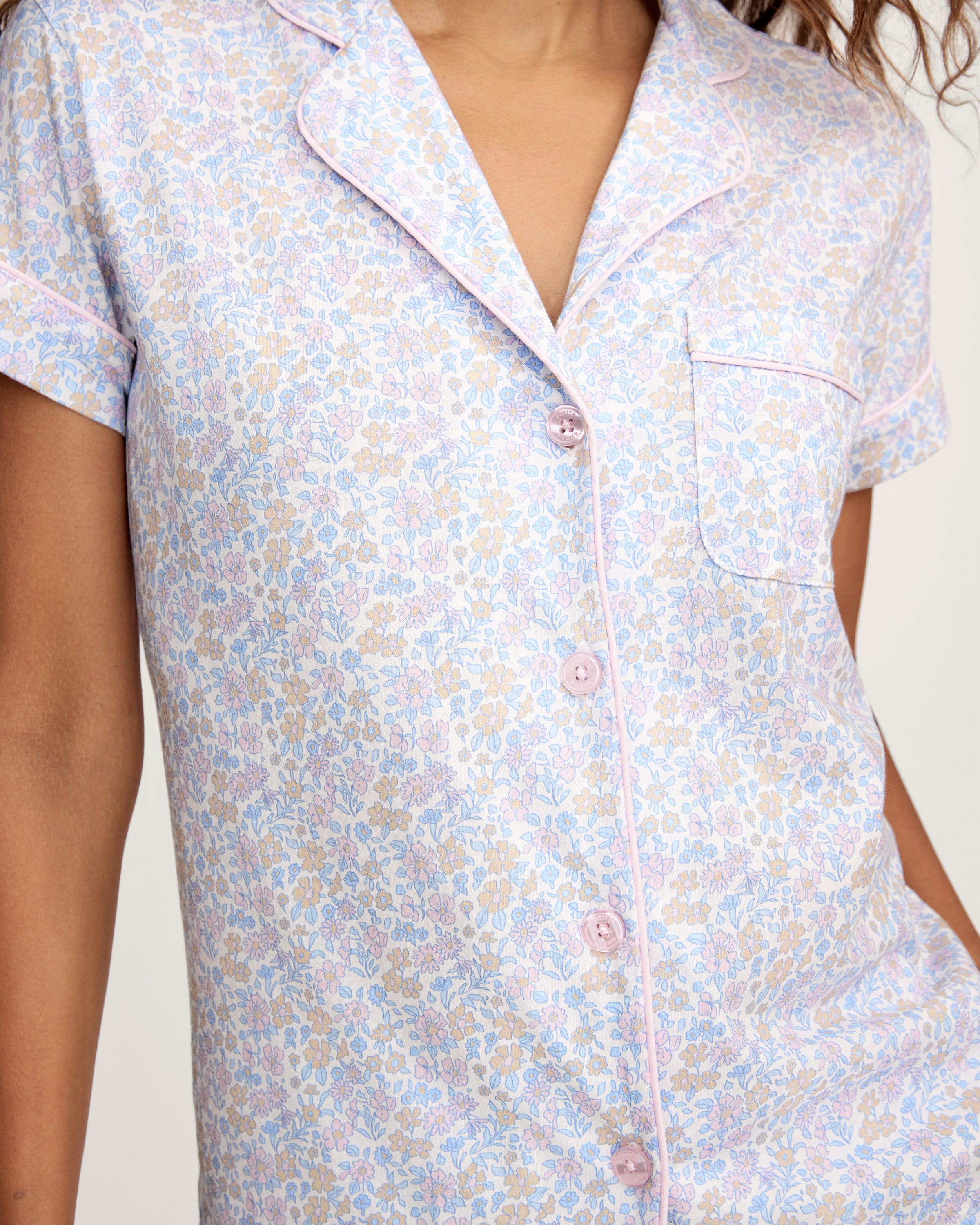 A woman with long wavy hair wears the Women's Lightweight Pima Short Sleeve Short Set in Fleur Blanche, featuring a pastel floral pattern, standing against a plain background.
