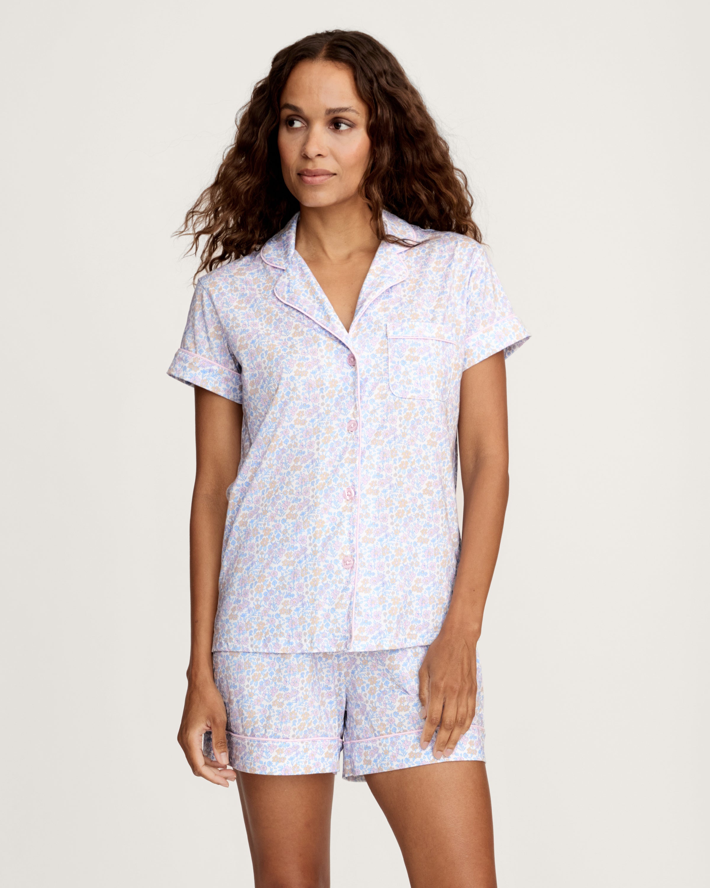 A woman with long wavy hair wears the Women's Lightweight Pima Short Sleeve Short Set in Fleur Blanche, featuring a pastel floral pattern, standing against a plain background.