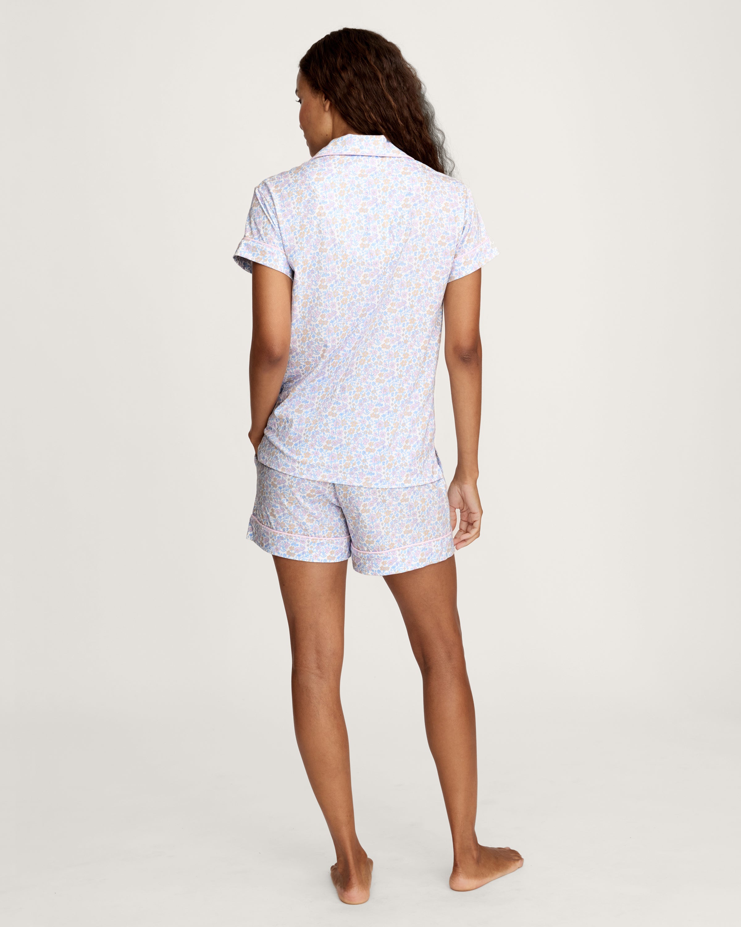 A woman with long wavy hair wears the Women's Lightweight Pima Short Sleeve Short Set in Fleur Blanche, featuring a pastel floral pattern, standing against a plain background.