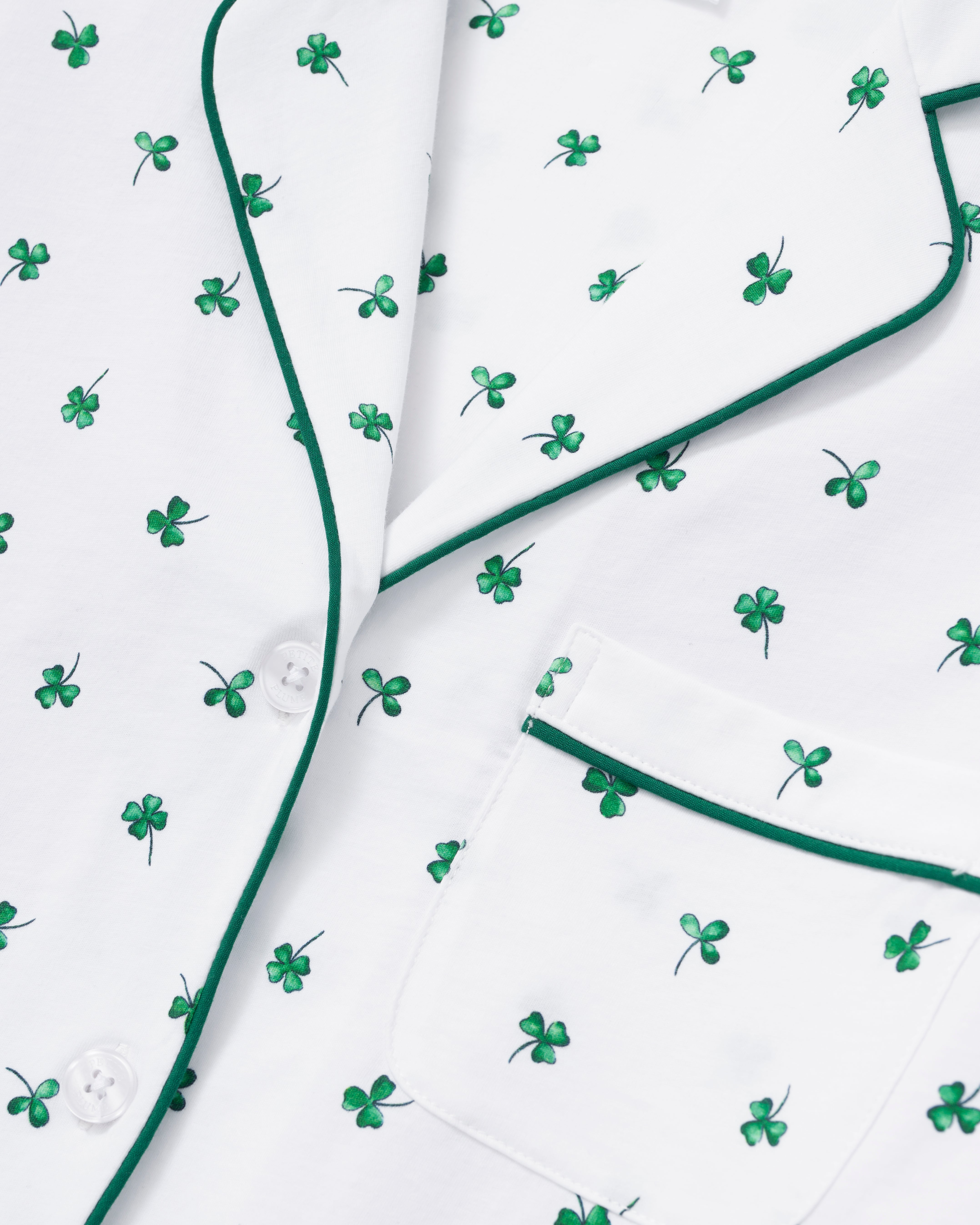 A woman with wavy blonde hair wears the Women's Pima Short Sleeve Short Set in Shamrocks—white pajamas with green clover prints, green trim, short-sleeve top and pocket, plus ruffled-hem shorts—standing against a plain background.