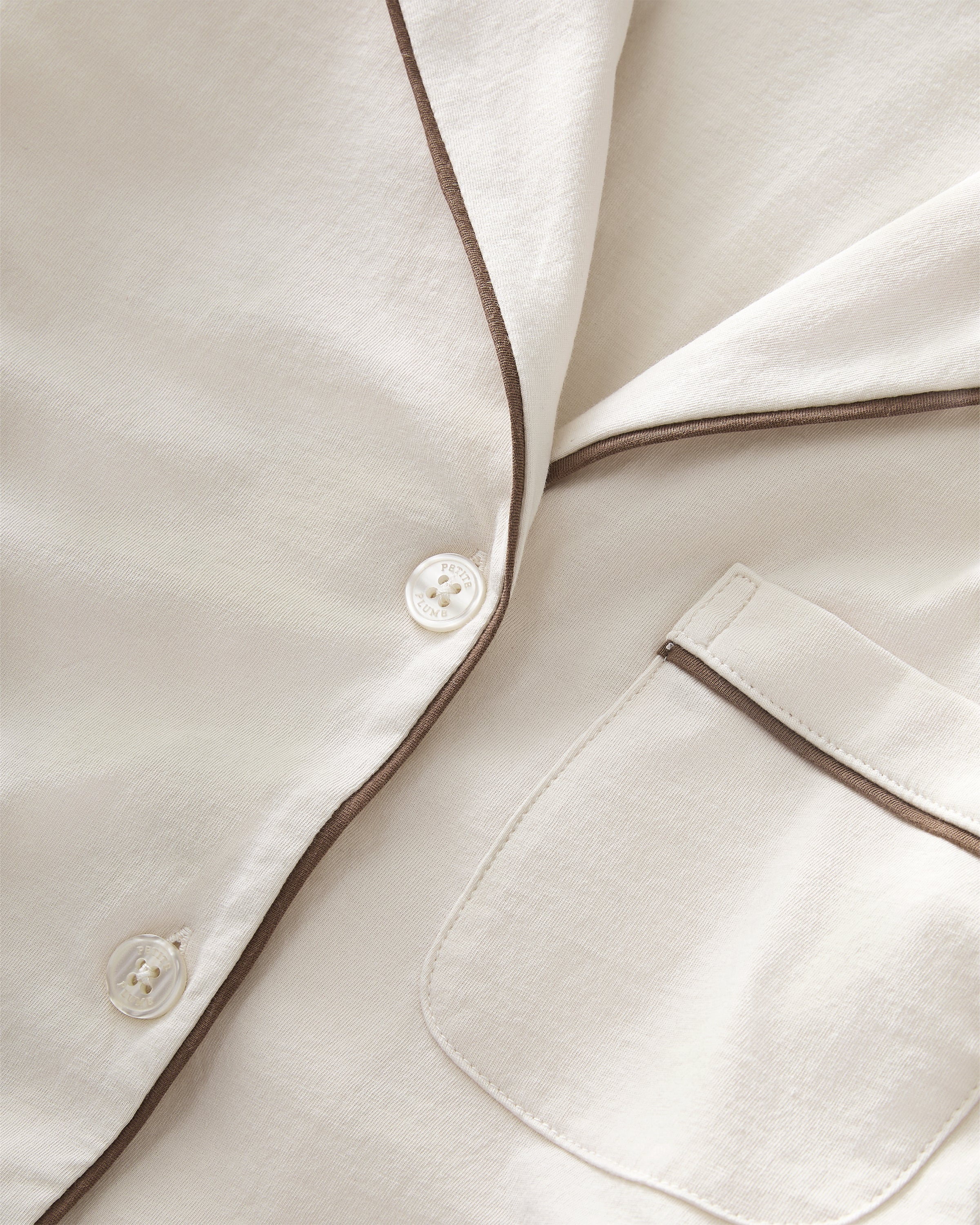 A woman with long curly hair models the Women's Lightweight Pima Short Sleeve Short Set in Ivory, featuring a button-up top with piping and matching shorts, standing barefoot against a plain light background.