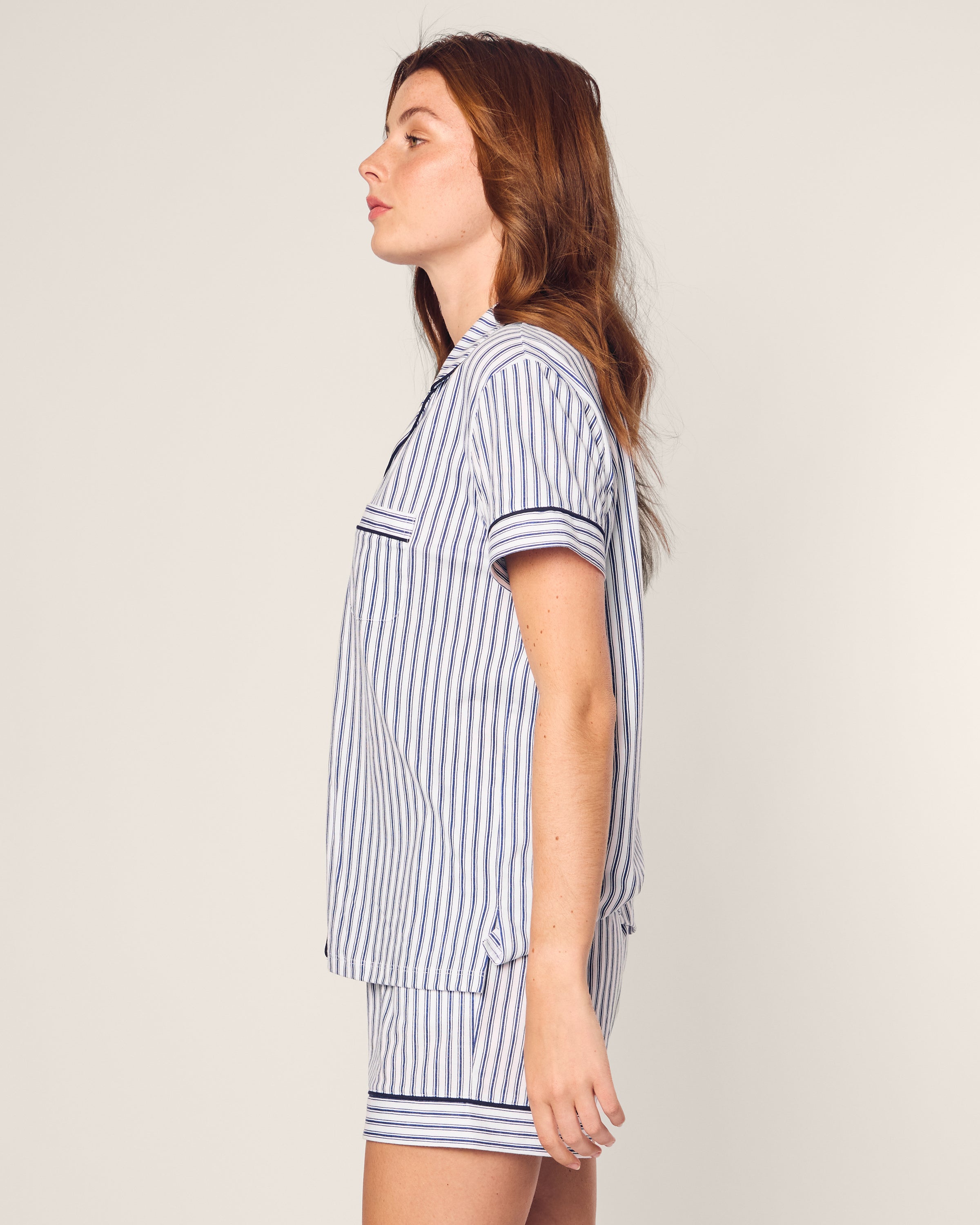 A woman stands in profile, showcasing the Womens Pima Pajama Short Set in Navy French Ticking by Petite Plume. She has long, loose hair and gazes straight ahead with an air of casual elegance against a plain off-white background.