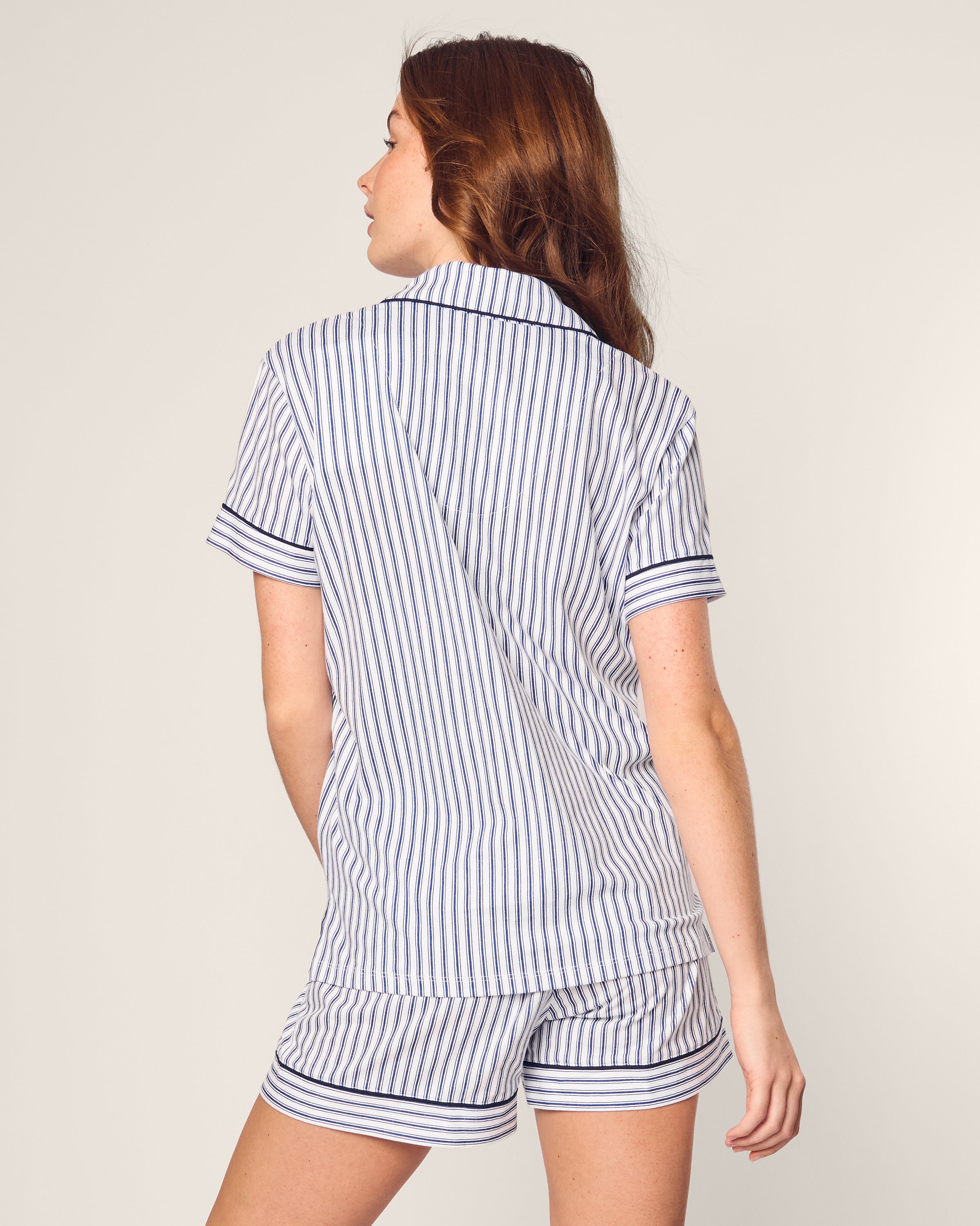 A woman wears the Petite Plume Womens Pima Pajama Short Set in Navy French Ticking. The cotton twill fabric showcases a blue and white stripe pattern with dark trim on the collar and cuffs, as she faces away against a light background.