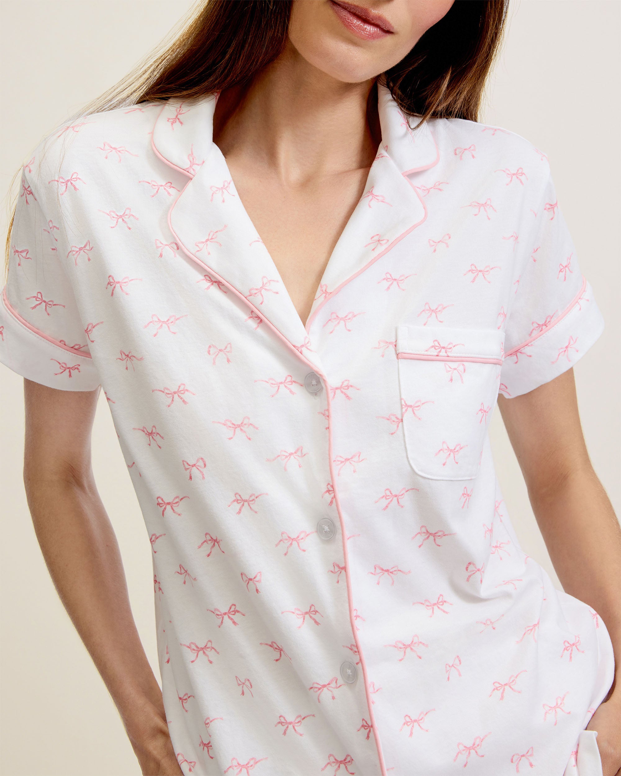 The Women's Pima Short Sleeve Short Set in Blushing Bows features a white button-up top with pink piping, a front pocket, and matching shorts, all adorned with an all-over pink bow pattern.
