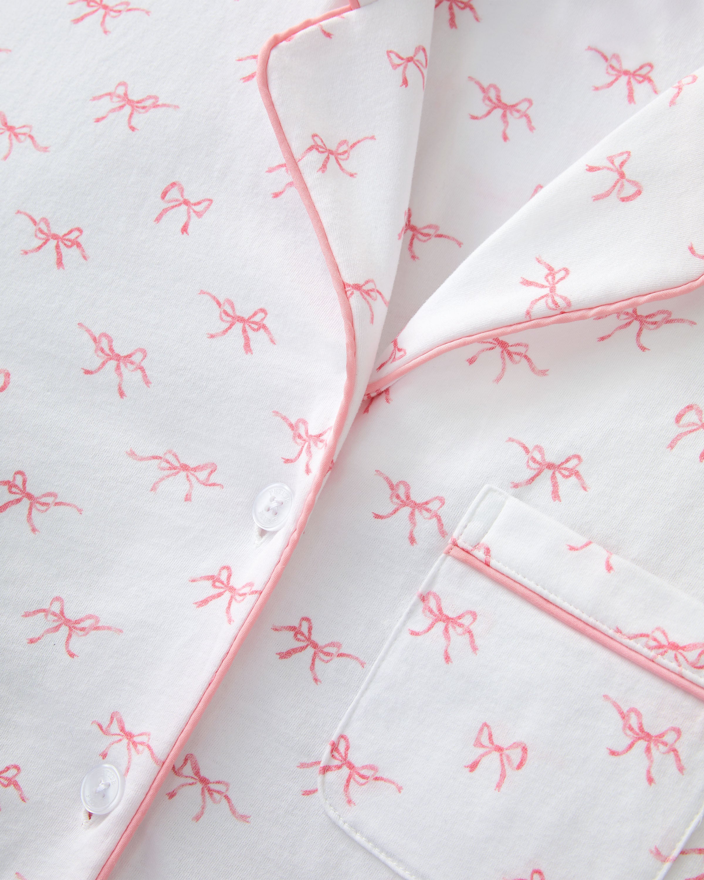 The Women's Pima Short Sleeve Short Set in Blushing Bows features a white button-up top with pink piping, a front pocket, and matching shorts, all adorned with an all-over pink bow pattern.