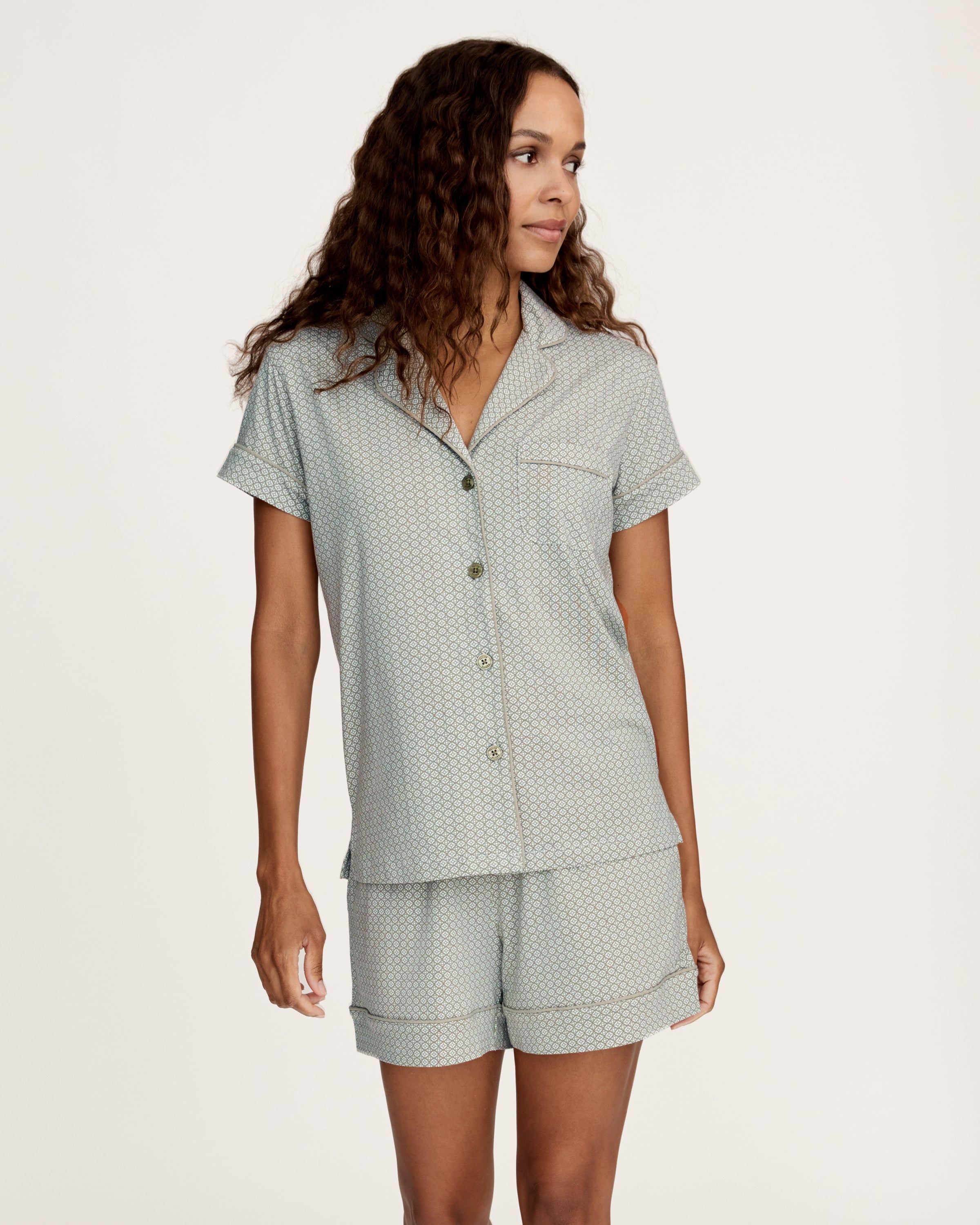 A woman stands barefoot in the Women's Lightweight Pima Short Sleeve Short Set in Sage Geo—featuring a button-up shirt and matching shorts with a small geometric pattern—against a plain light background.