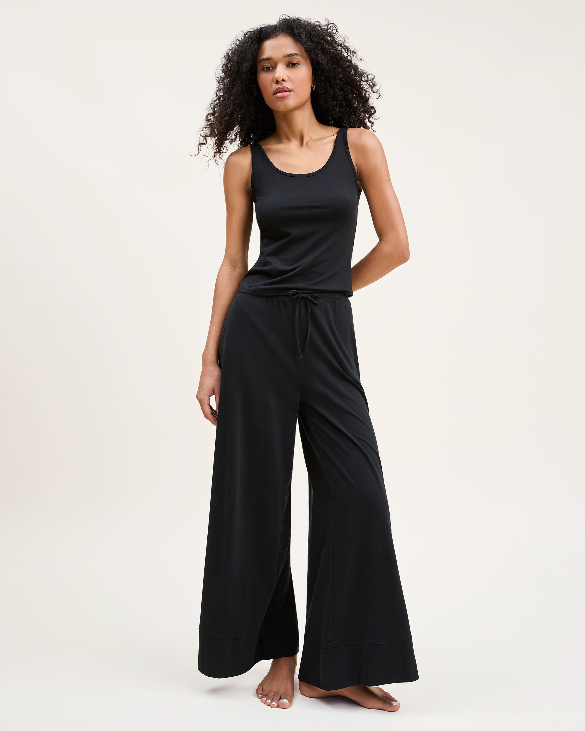 A woman with curly hair stands barefoot against a plain background, wearing the Women's Pima Wide-Leg Lounge Pant in Black and a sleeveless black tank top, looking relaxed and confident.
