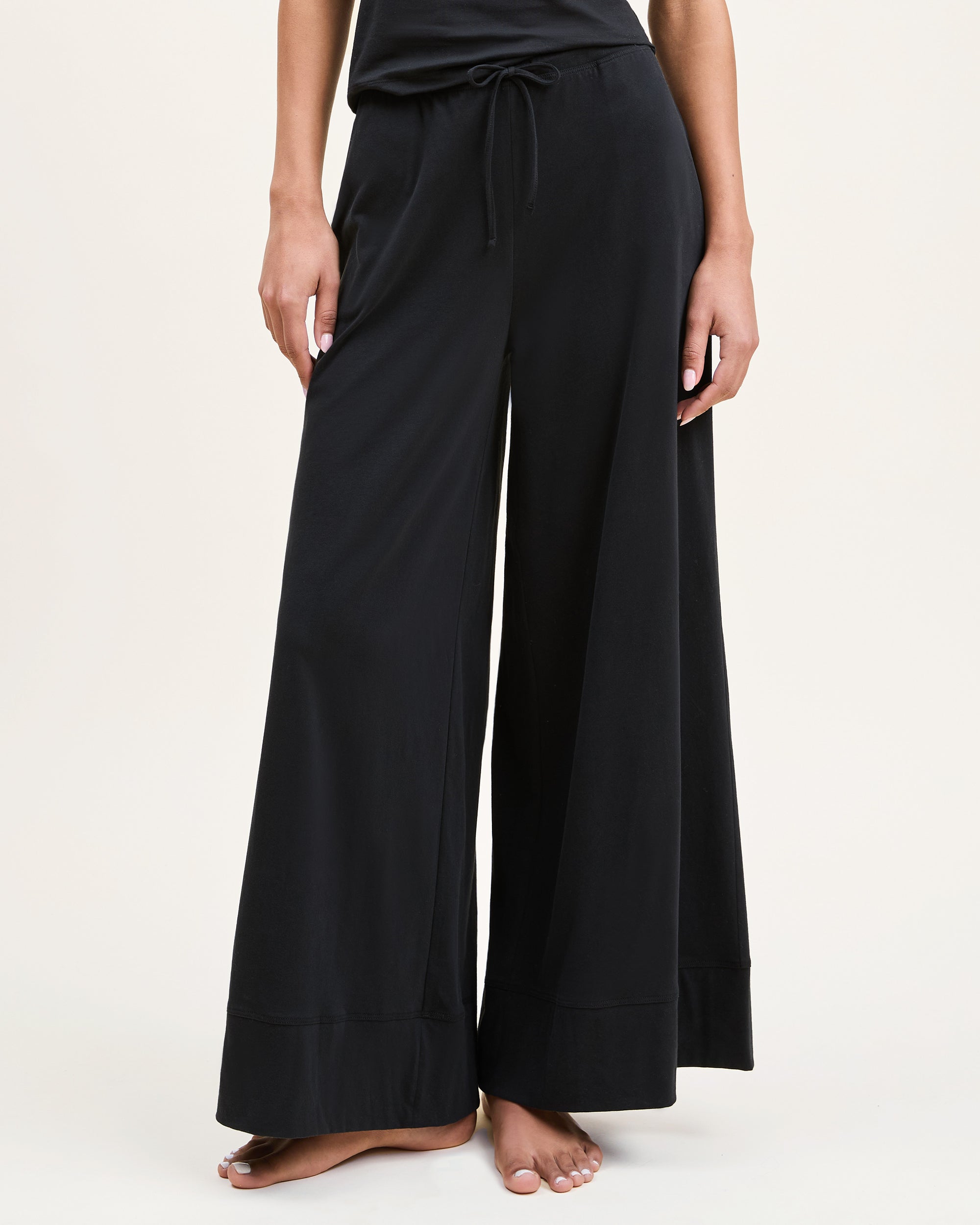 A woman with curly hair stands barefoot against a plain background, wearing the Women's Pima Wide-Leg Lounge Pant in Black and a sleeveless black tank top, looking relaxed and confident.