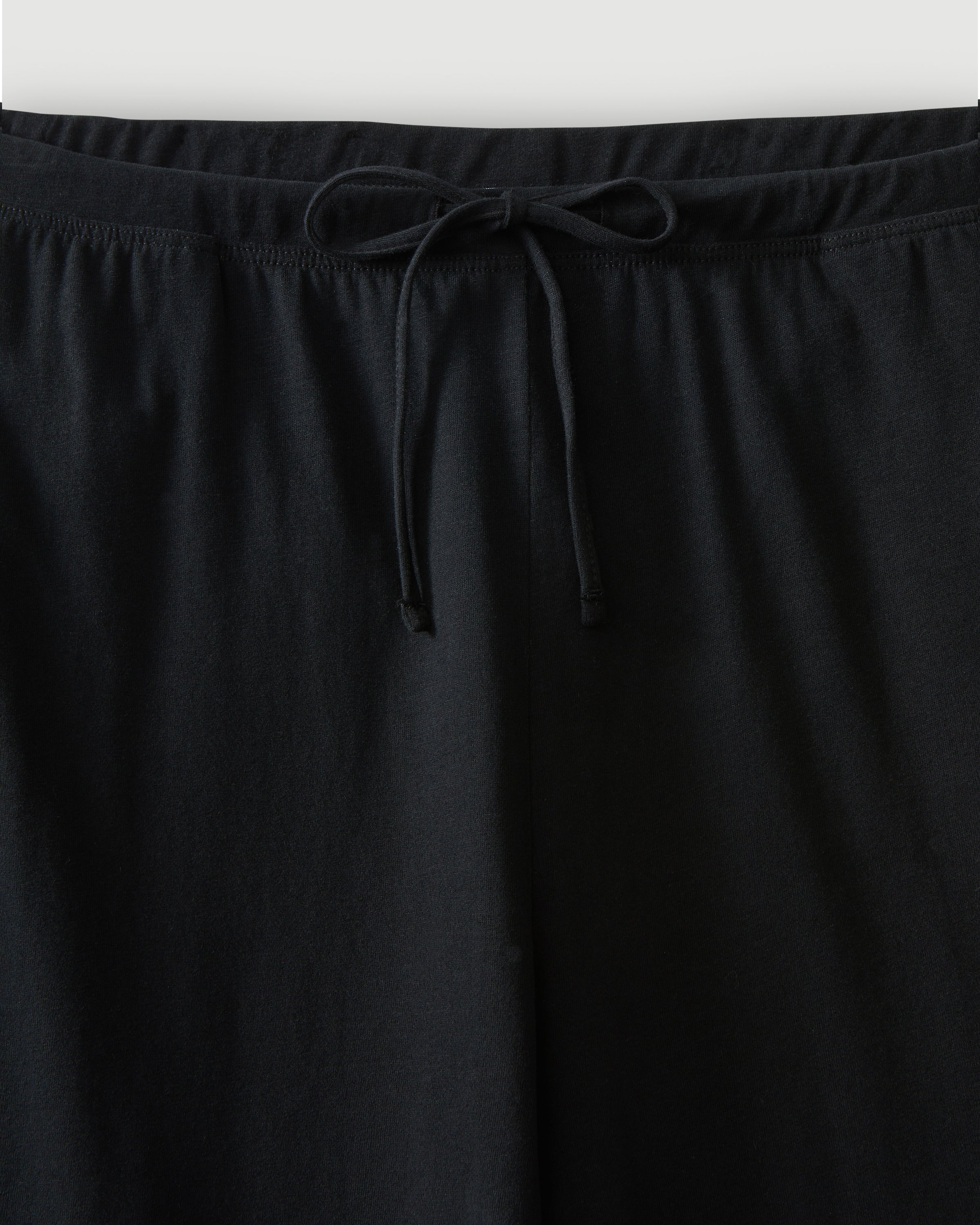 A woman with curly hair stands barefoot against a plain background, wearing the Women's Pima Wide-Leg Lounge Pant in Black and a sleeveless black tank top, looking relaxed and confident.