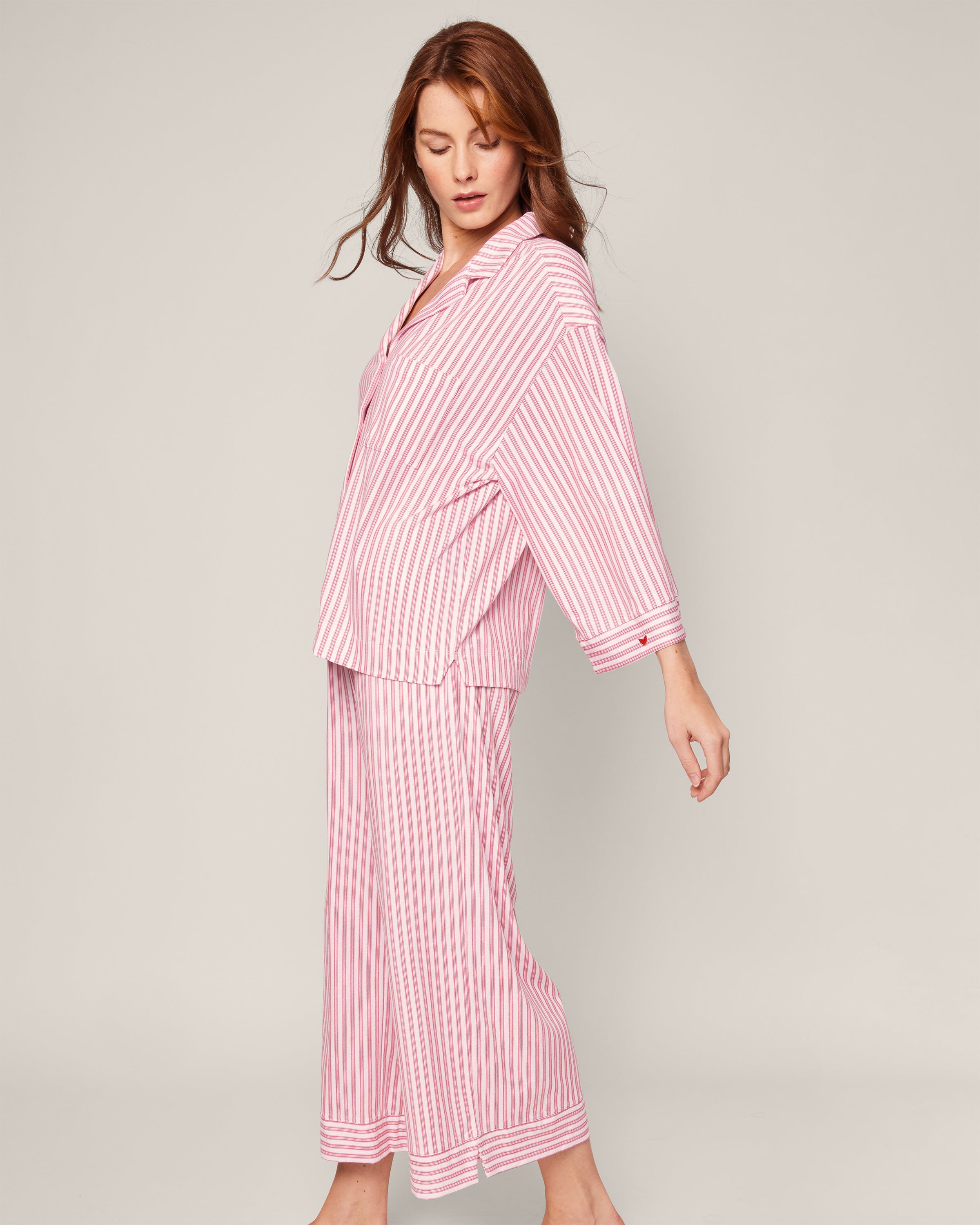 A person with long brown hair stands against a plain backdrop, looking slightly down to the right with one leg bent, wearing Petite Plumes loose-fitting and comfy Olivia Wide Leg Pima Pajama Set in Antique Red Ticking.
