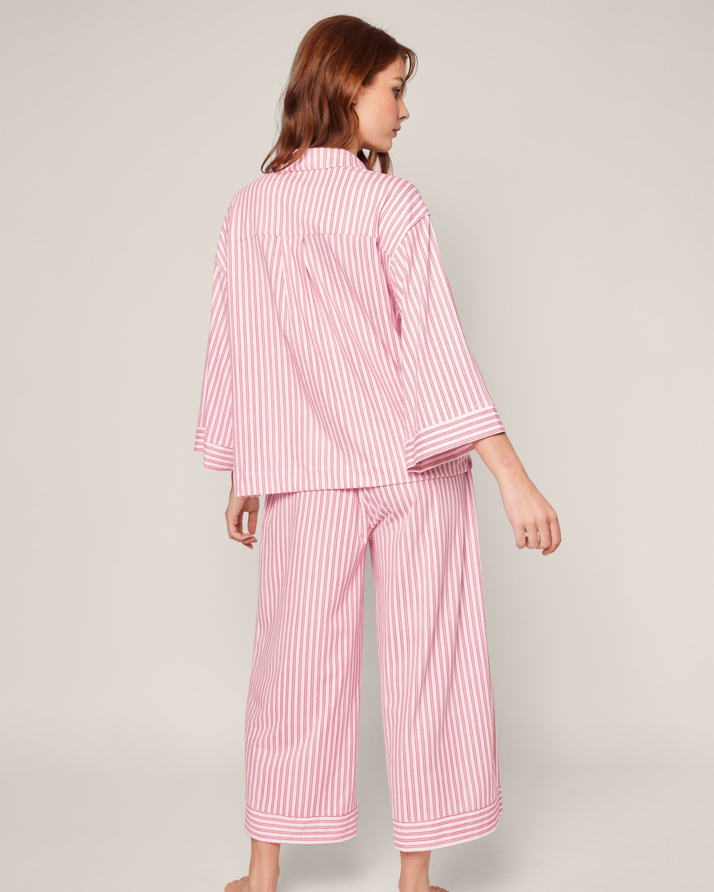 A person with long hair is barefoot on a plain background, wearing Petite Plumes The Olivia Wide Leg Pima Pajama Set in Antique Red Ticking, showcasing the back and side of the loose-fitting pajamas as they face slightly away from the camera.