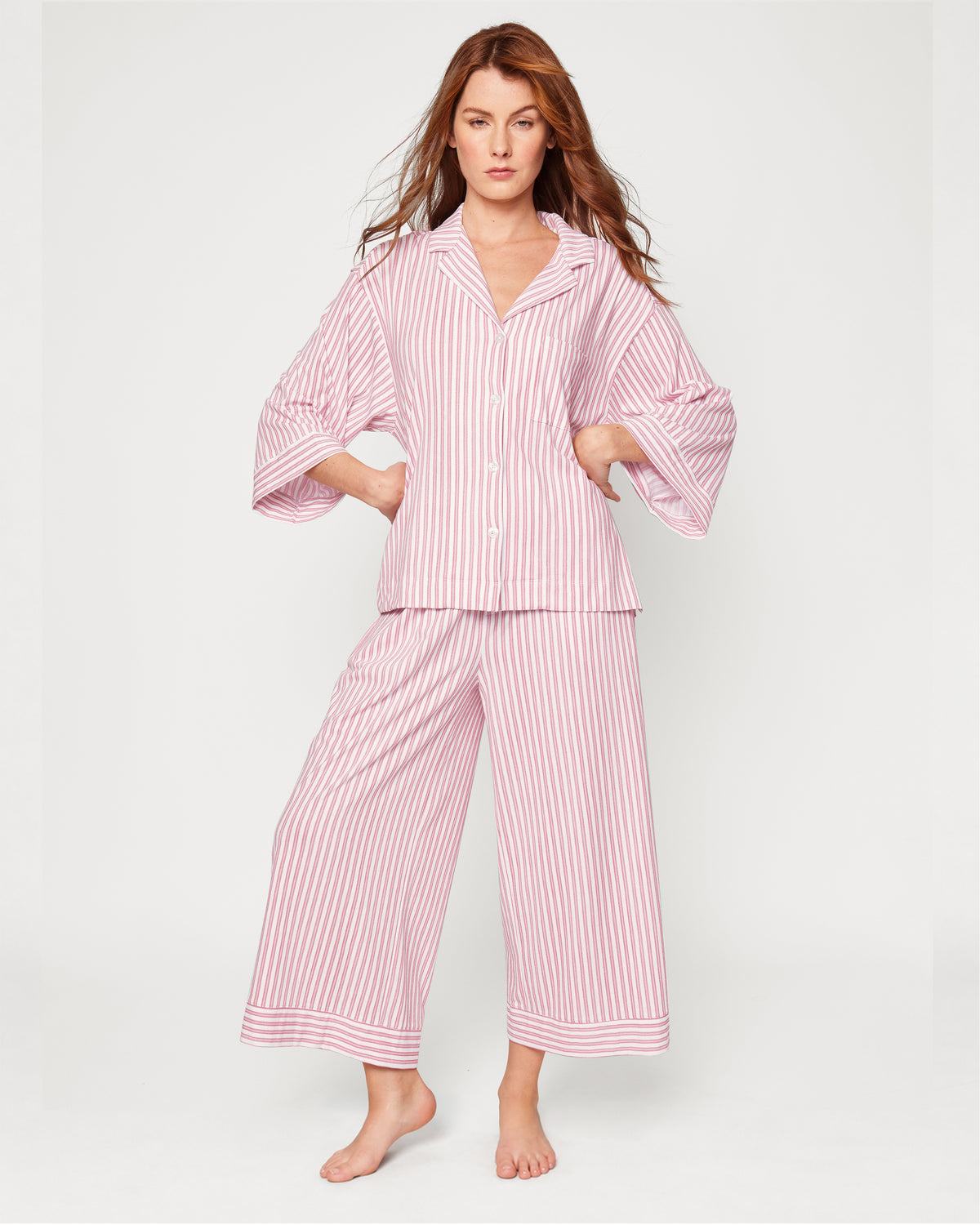 A person wearing The Olivia Wide Leg Pima Pajama Set in Antique Red Ticking, featuring a long-sleeved button-up shirt and wide-leg trousers in a red and white striped pattern.
