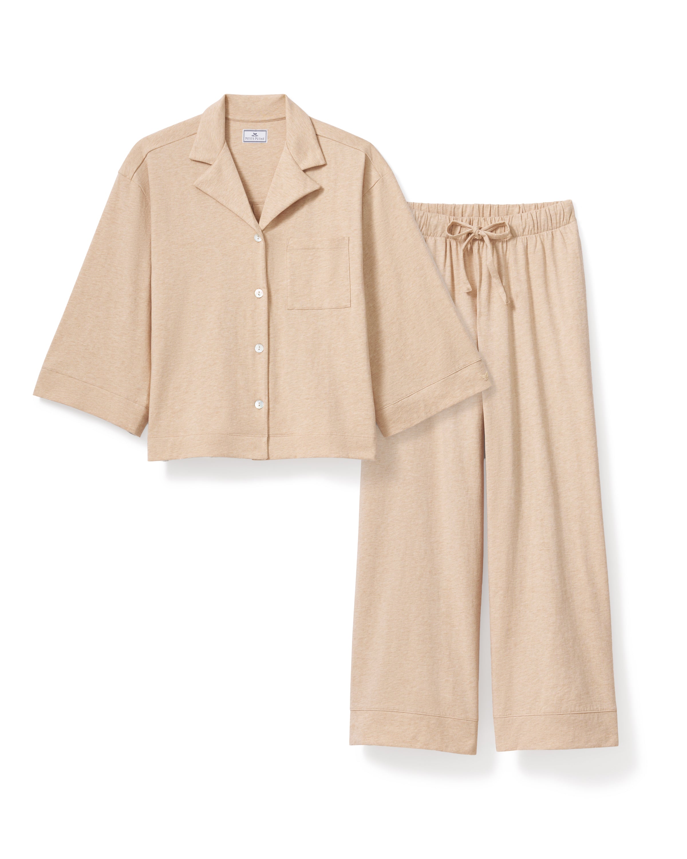 Against a plain backdrop, an individual with long curls and a relaxed stance wears The Olivia Wide Leg Pima Pajama Set in Camel by Petite Plume. The luxurious set features a button-up top and loose-fit pants, crafted from soft Pima cotton. They are barefoot, embodying comfort and elegance.