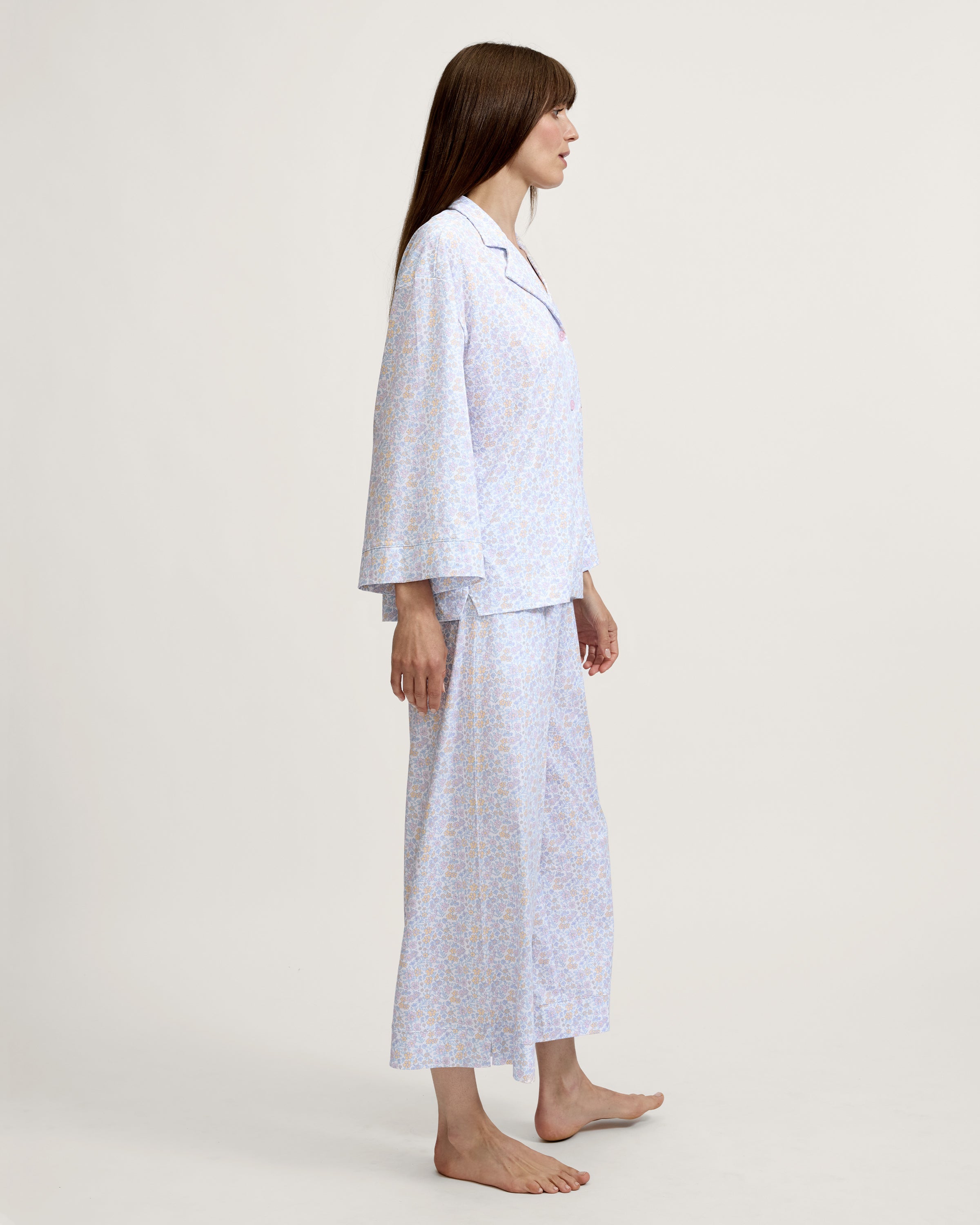 A woman stands barefoot against a plain background, wearing The Olivia Wide Leg Lightweight Pima Pajama Set in Fleur Blanche, featuring a loose long-sleeve button-up top and wide-leg pants with a light pastel floral pattern. She looks to the side and smiles.