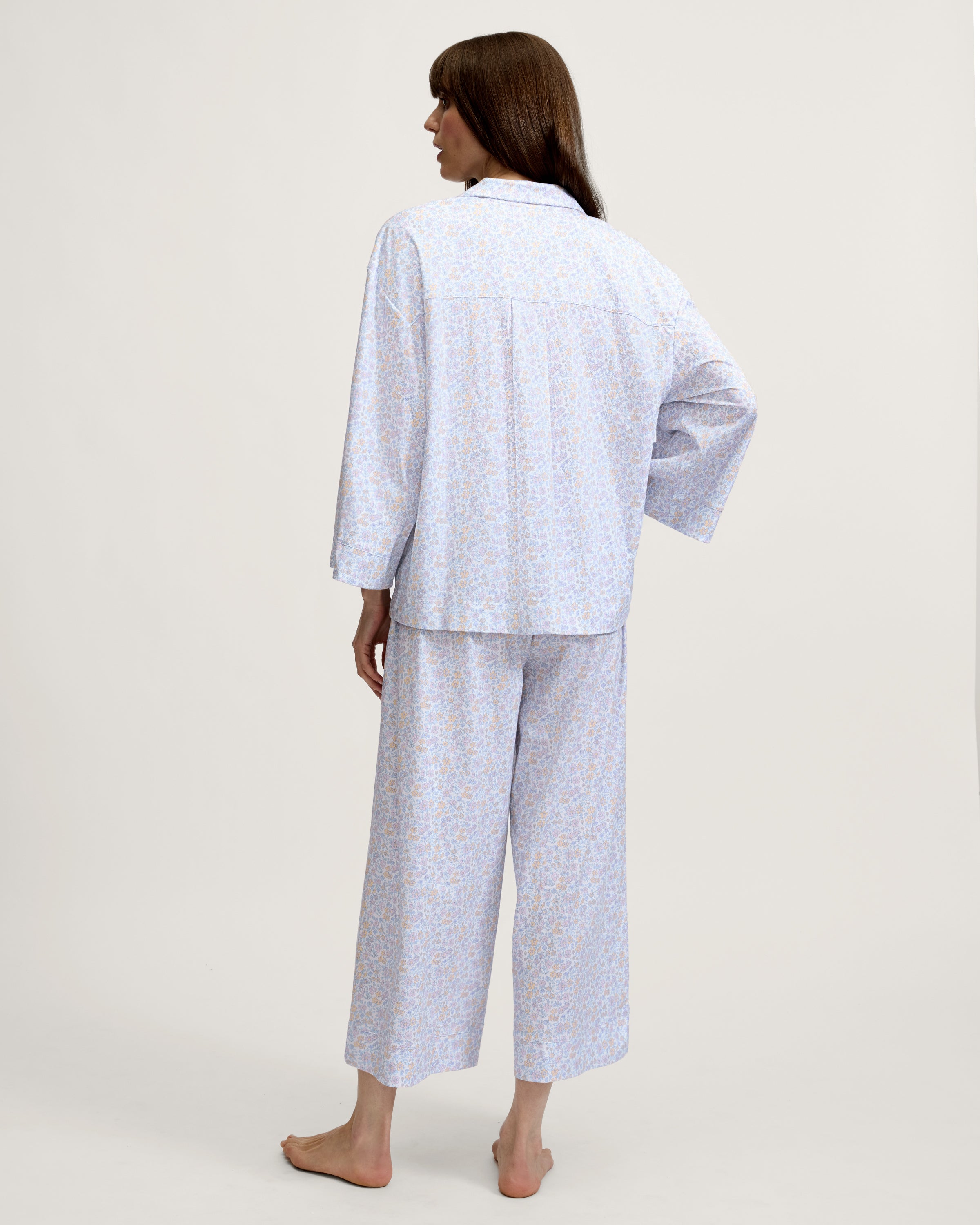 A woman stands barefoot against a plain background, wearing The Olivia Wide Leg Lightweight Pima Pajama Set in Fleur Blanche, featuring a loose long-sleeve button-up top and wide-leg pants with a light pastel floral pattern. She looks to the side and smiles.