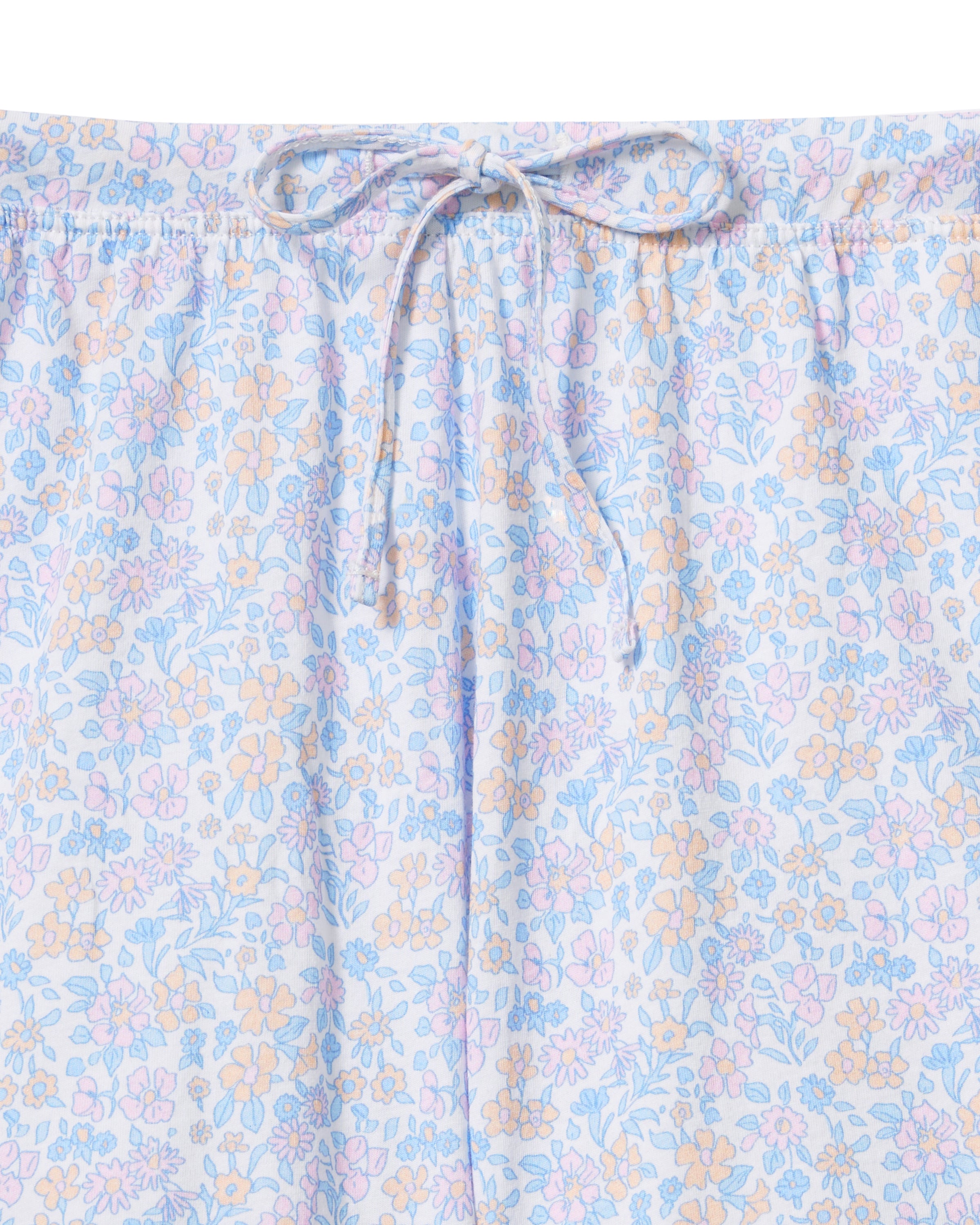 A woman stands barefoot against a plain background, wearing The Olivia Wide Leg Lightweight Pima Pajama Set in Fleur Blanche, featuring a loose long-sleeve button-up top and wide-leg pants with a light pastel floral pattern. She looks to the side and smiles.