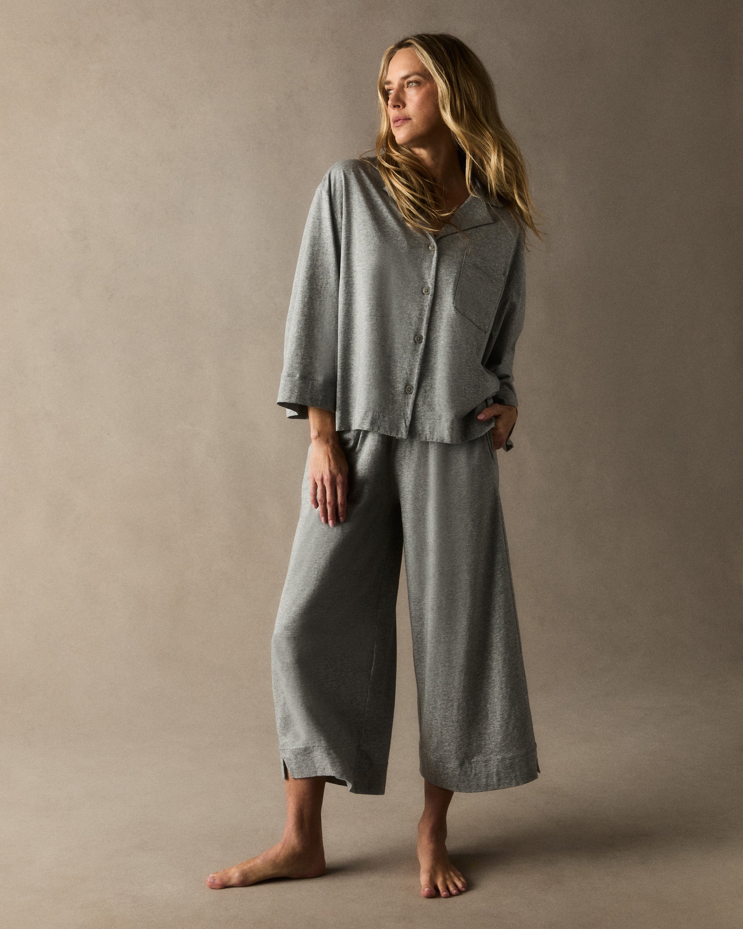 A woman stands barefoot against a plain background, smiling gently with one hand by her side, wearing The Olivia Wide Leg Lightweight Pima Pajama Set in Heather Grey—loose-fitting pants and a matching long-sleeve button-up top.