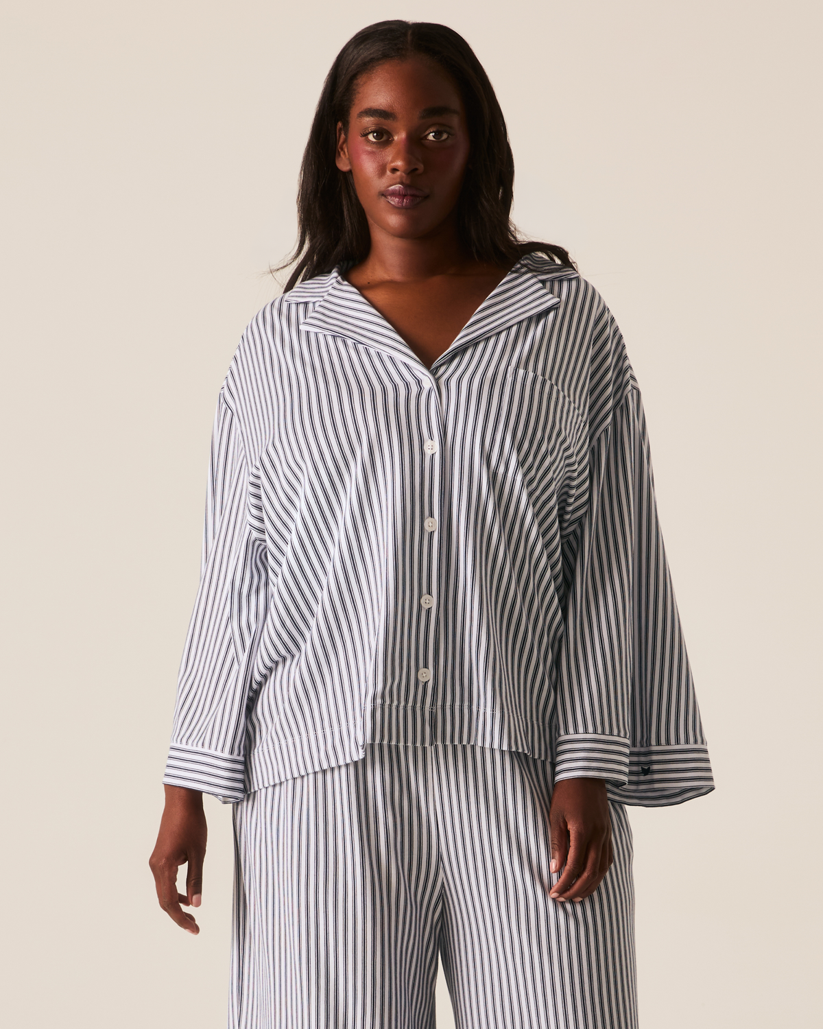 A woman wears The Olivia Wide Leg Pima Pajama Set in Navy French Ticking, featuring a long-sleeved button-down shirt and wide-leg pants in a navy and white pinstripe pattern.