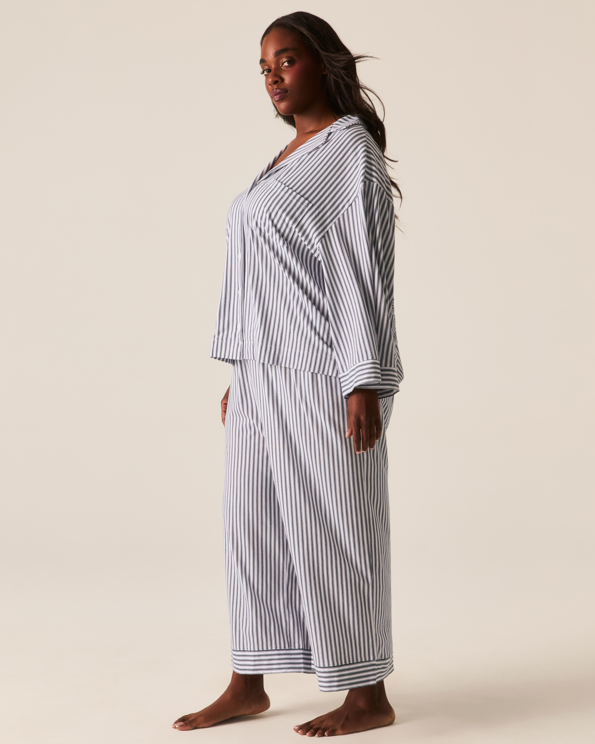 A person wearing The Olivia Wide Leg Pima Pajama Set in Navy French Ticking, featuring a long-sleeved, button-down top and matching wide-leg trousers in a blue and white pinstripe pattern.