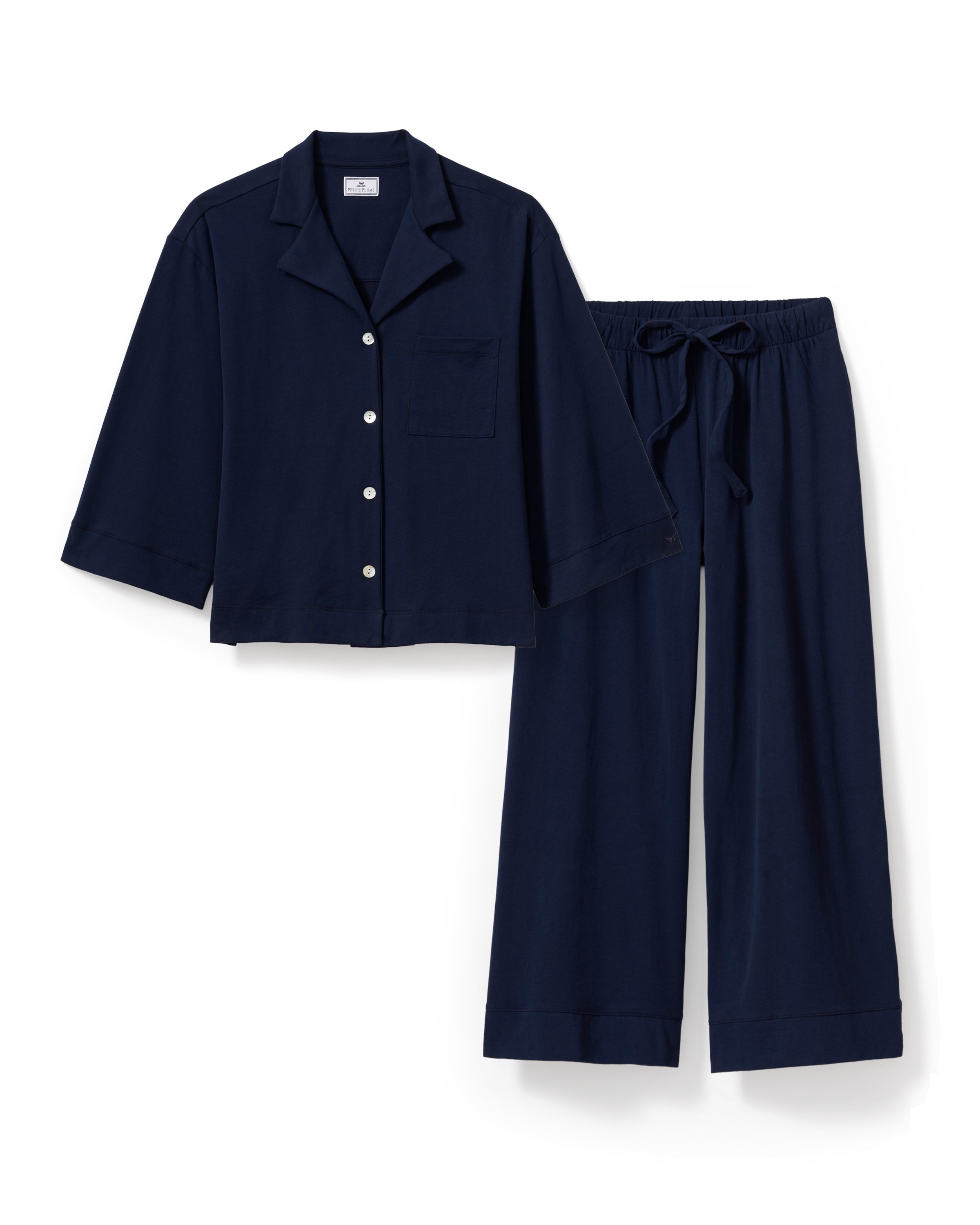A person with long brown hair wears The Olivia Wide Leg Pima Pajama Set in Navy by Petite Plume, featuring a luxurious, buttoned, long-sleeved top and elegant wide-legged pants. They stand barefoot against an off-white background.